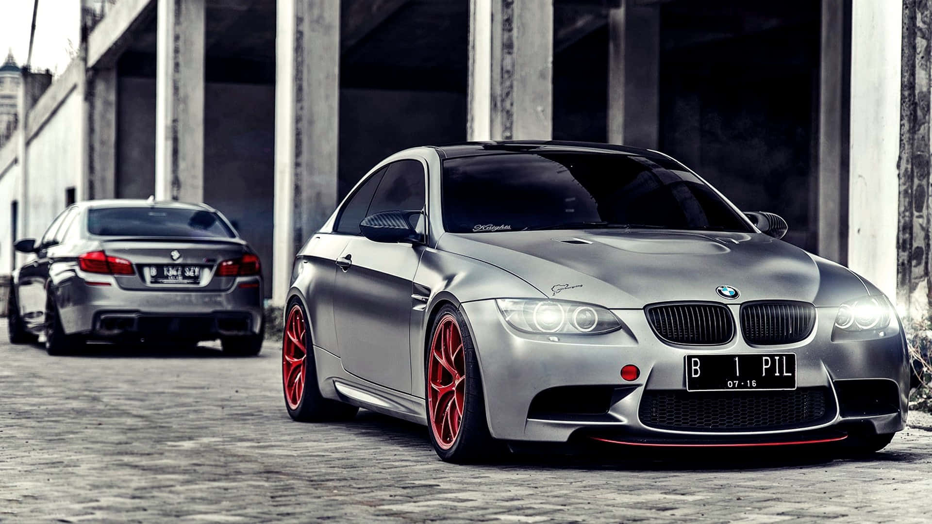 1920x1080 Bmw 桌布 壁紙 — 一輛時尚的銀色BMW M3配以鮮紅色輪圈，停在混凝土柱下的鵝卵石街道上，後面還有另一輛車。