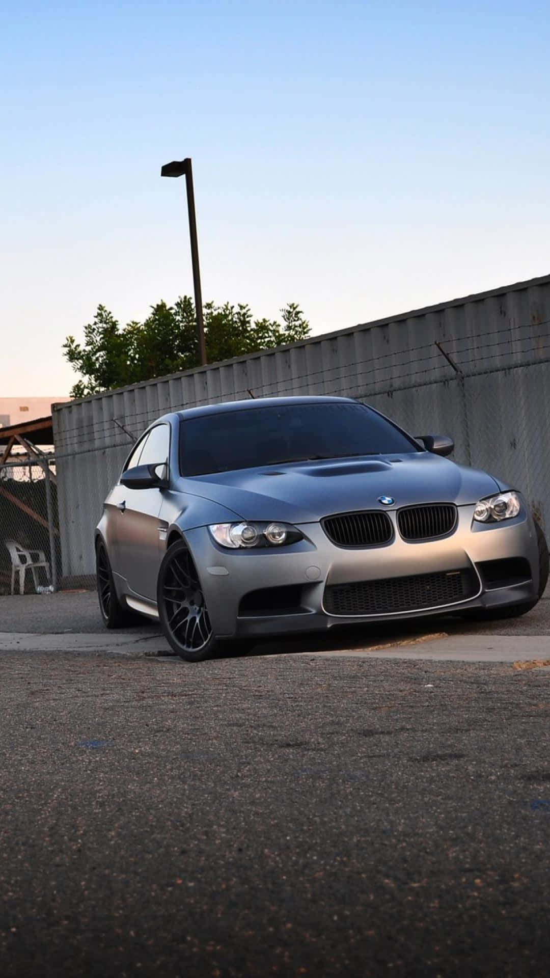 Bmw Android 桌布 壁紙 — 一輛時尚的銀色BMW M3雙門跑車停在瀝青路上，旁邊有鐵絲網圍欄和樹木，在黃昏時分清澈的藍天下。