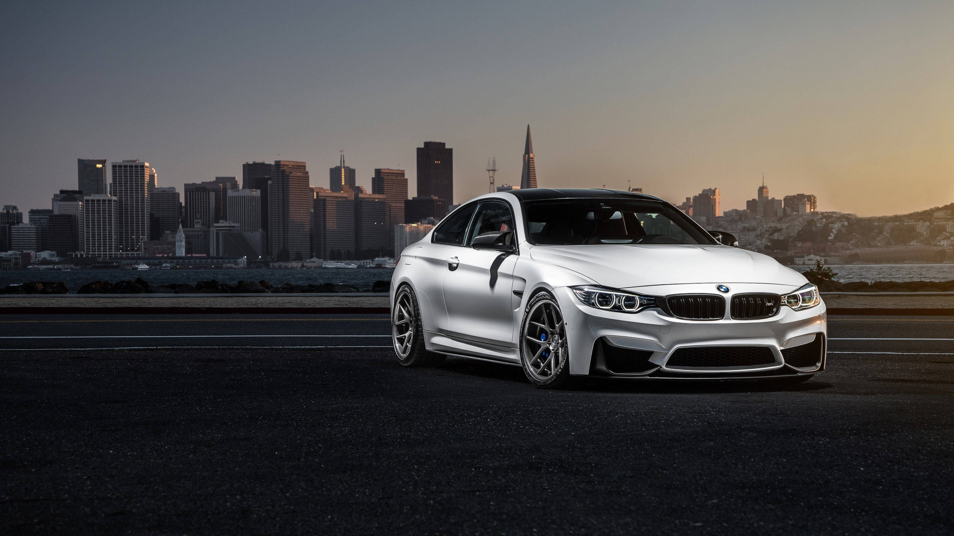 4K BMW 壁紙 桌布 壁紙 — 一輛時尚的銀色BMW M4轎跑車停在高速公路上，舊金山的天際線在日落時分閃耀著光芒。