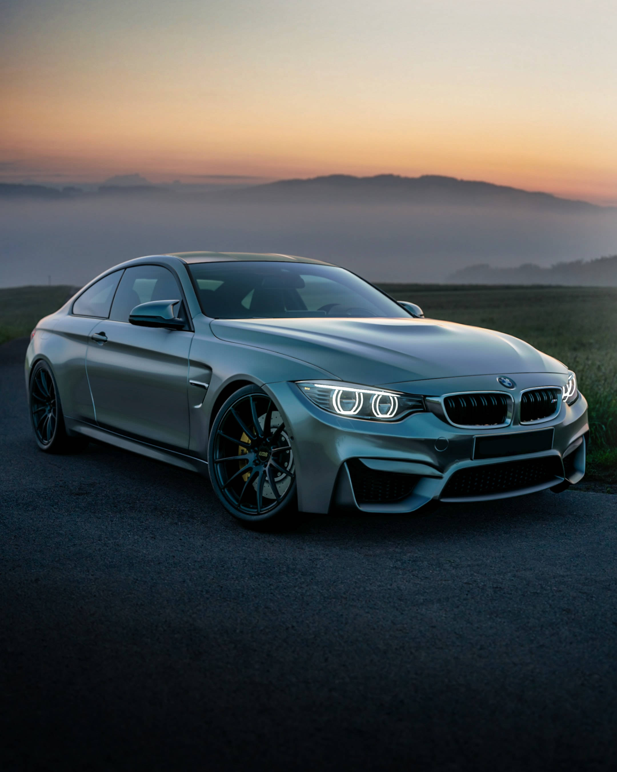 BMW 動圖 桌布 壁紙 — 一輛時尚的銀色BMW M4雙門跑車在黎明時分於霧氣瀰漫的山路上閃耀，車頭燈亮起。