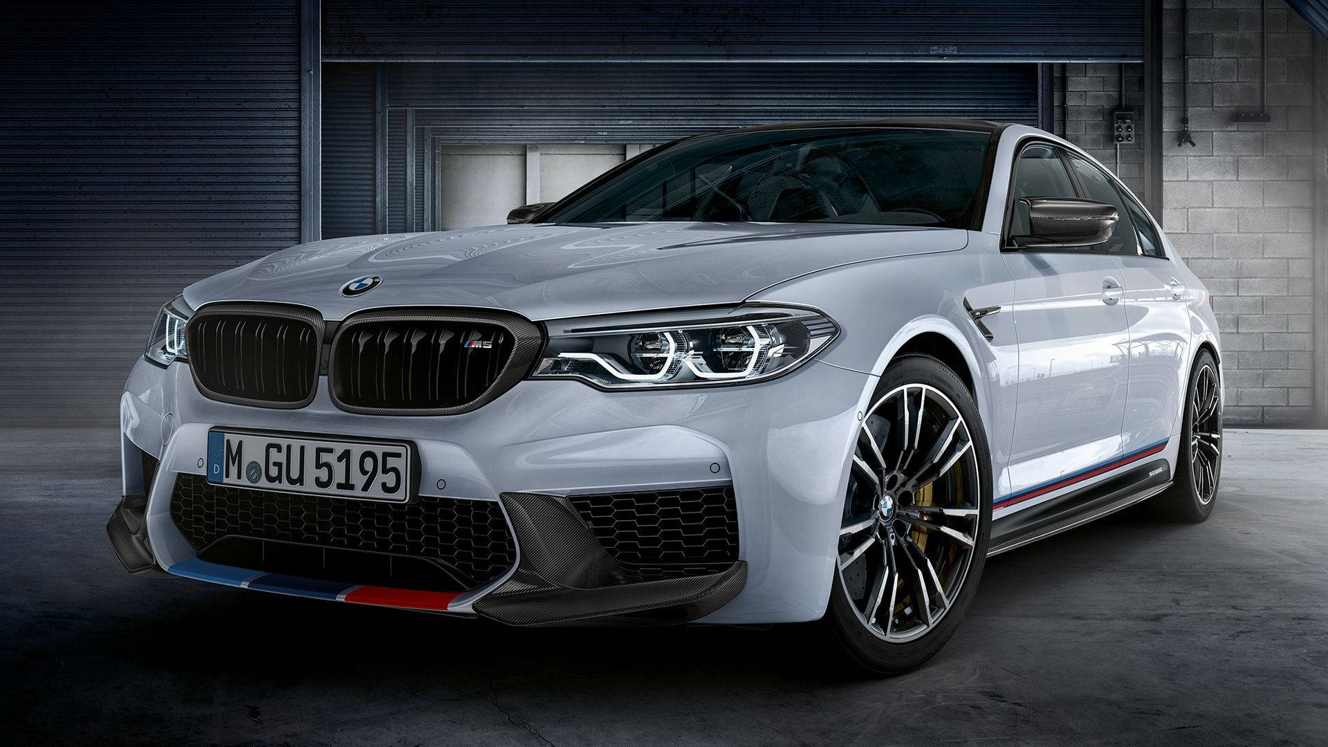 m 桌布 壁紙 — 一輛時尚的銀色BMW M5轎車，帶有紅、藍、白賽車條紋，停放在光線昏暗的工業車庫中。