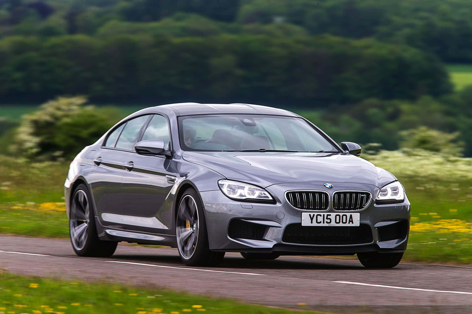 Bmw 6 Series 桌布 壁紙 — 一輛時尚的銀色BMW M6雙門跑車在蜿蜒的鄉村道路上飛馳，周圍環繞著鬱鬱蔥蔥的綠色山丘和野花。