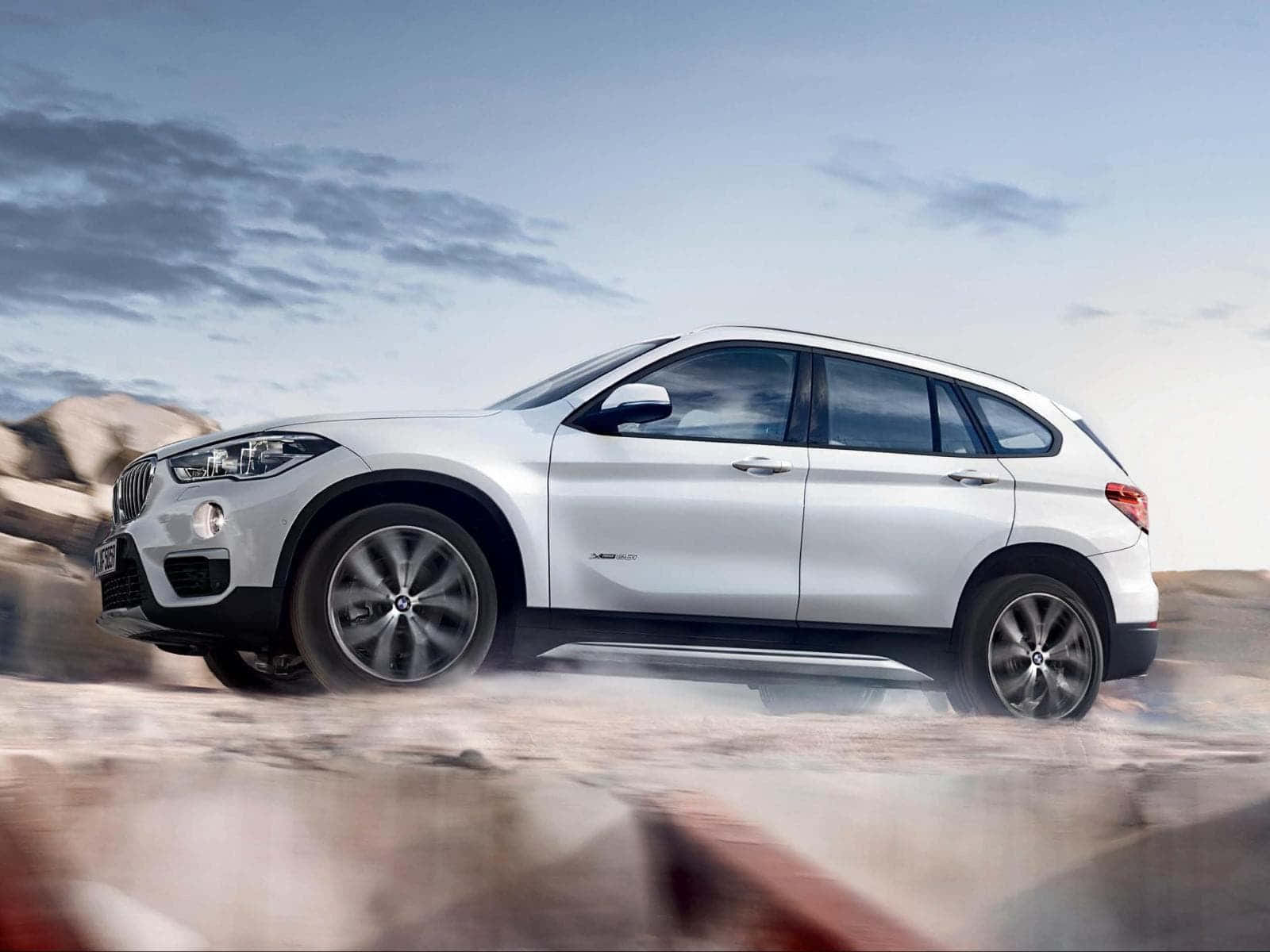 Bmw I8 桌布 壁紙 — 一輛時尚的銀色BMW X1 SUV在多雲的天空下，在塵土飛揚的沙漠景觀中疾馳。