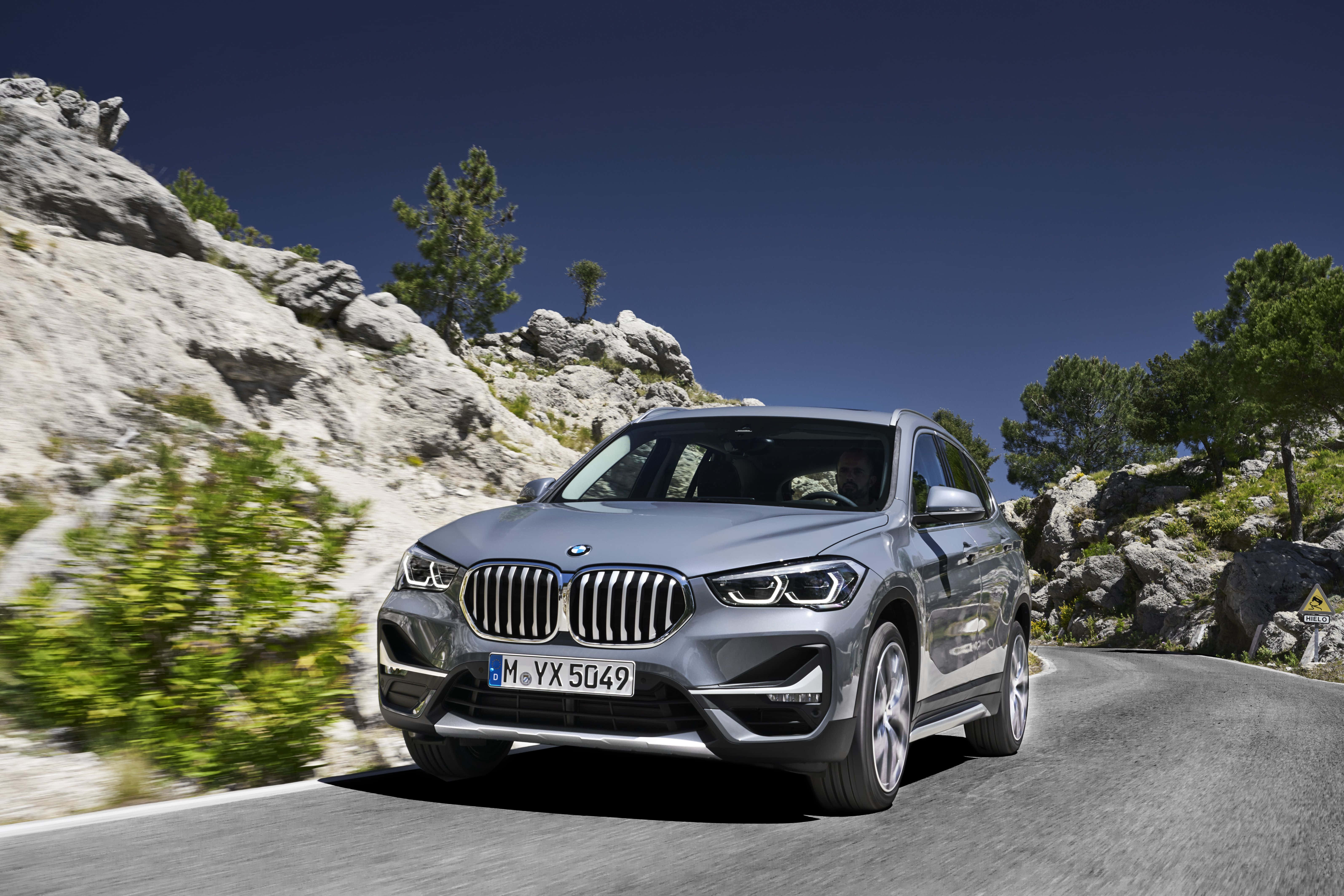 Bmw I8 桌布 壁紙 — 一輛時尚的銀色BMW X1 SUV在晴朗的藍天下，沿著蜿蜒的山路行駛，兩旁是岩石峭壁和綠樹。