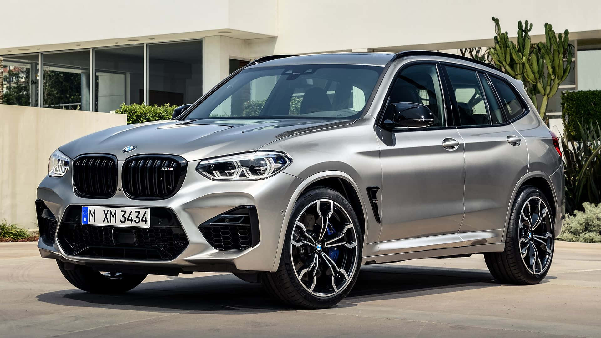 Bmw X3 桌布 壁紙 — 一輛時尚的銀色BMW X3 M停泊在陽光普照的車道上，旁邊是一座有仙人掌和綠植的現代房屋。