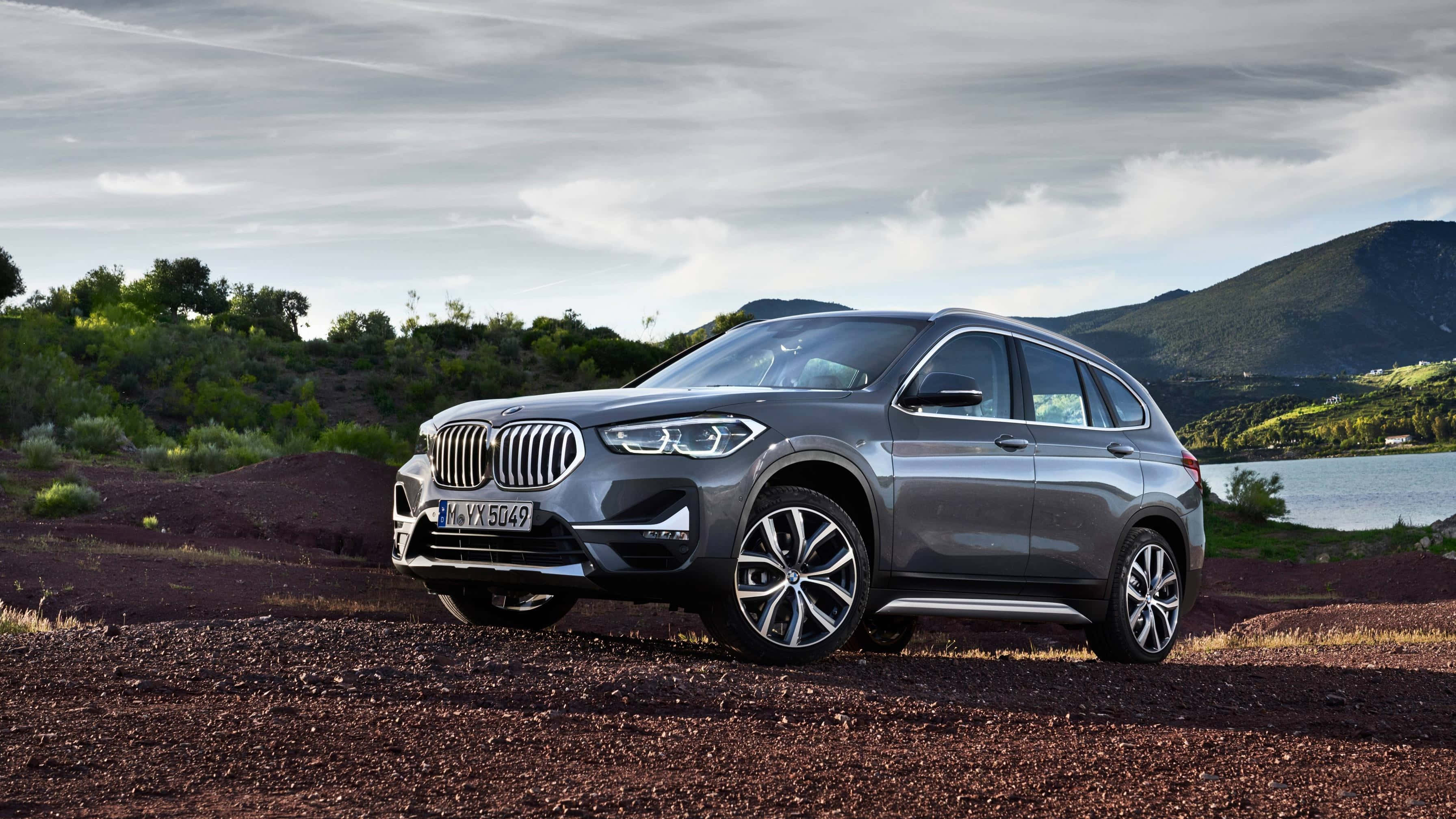 Bmw X1 桌布 壁紙 — 一輛時尚的銀色BMW X3 SUV停泊在平靜湖泊旁的紅褐色泥路上，背景是綠丘和陰天。