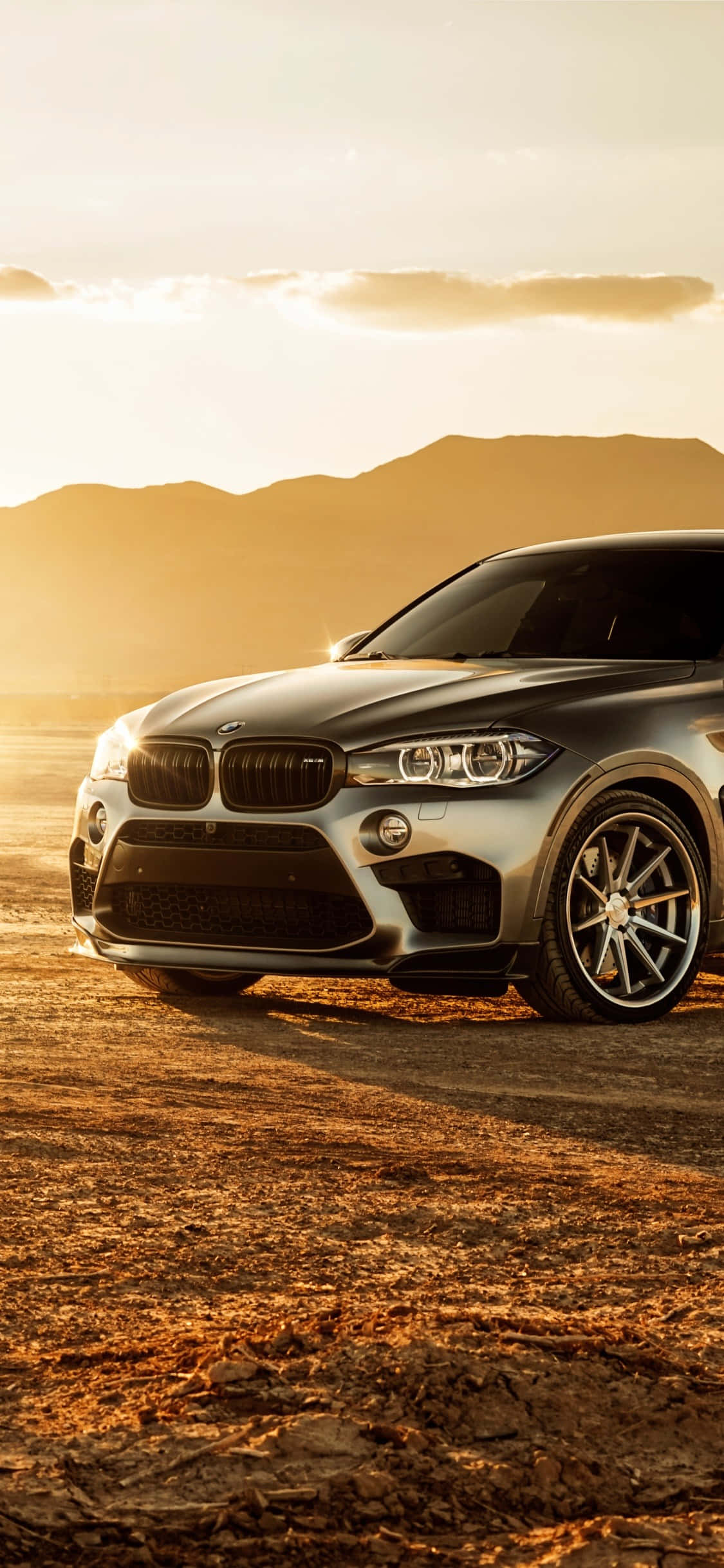 iPhone X Bmw 桌布 壁紙 — 一輛時尚的銀色BMW X6 SUV停放在塵土飛揚的地形上，背景是金色夕陽和遠山。