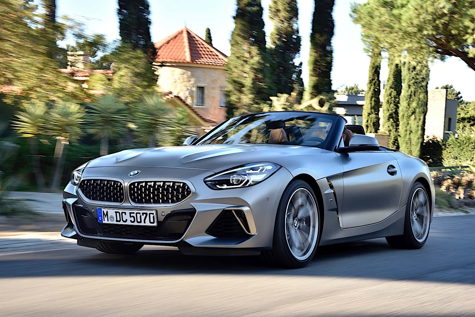 Bmw Z4 桌布 壁紙 — 一輛時尚的銀色BMW Z4敞篷跑車在陽光明媚的道路上奔馳，兩旁種滿高大的柏樹和地中海風格的房屋。