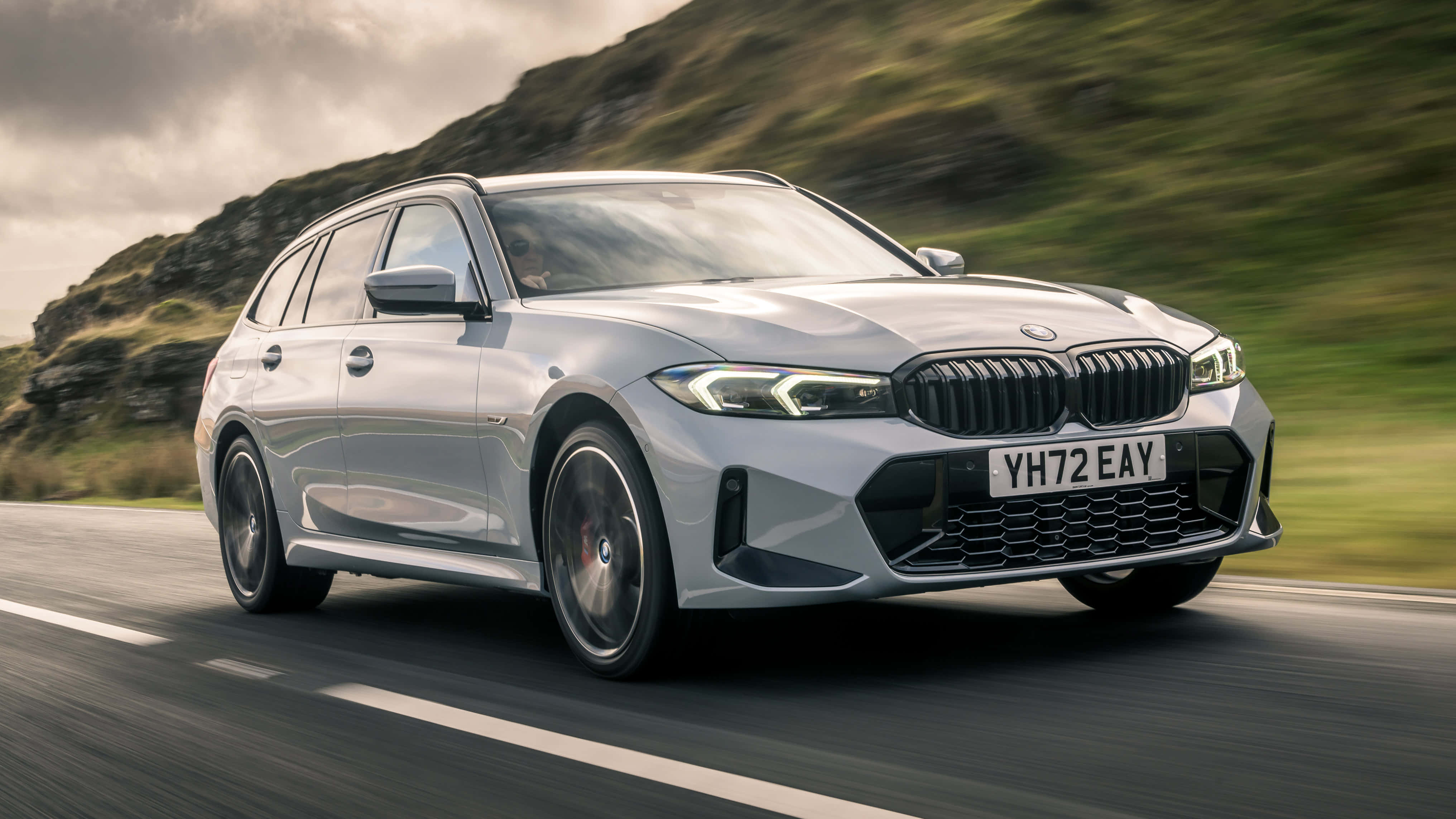 Bmw 3 Series 桌布 壁紙 — 一輛時尚的銀色BMW旅行車沿著蜿蜒的道路疾馳，背景是模糊的綠色山丘。