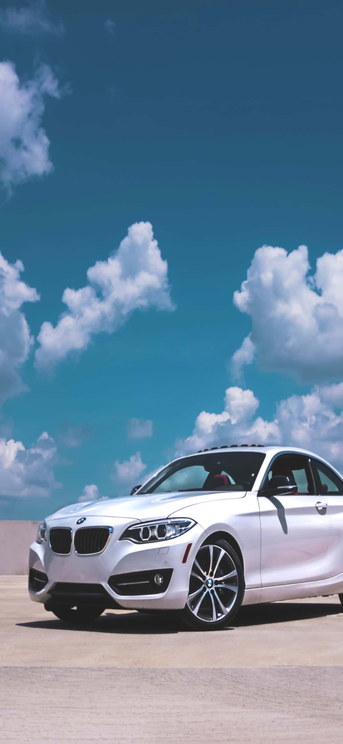 iPhone X Bmw 桌布 壁紙 — 一輛時尚的銀色BMW雙門轎跑車停在混凝土上，上方是充滿蓬鬆白雲的鮮豔藍天。