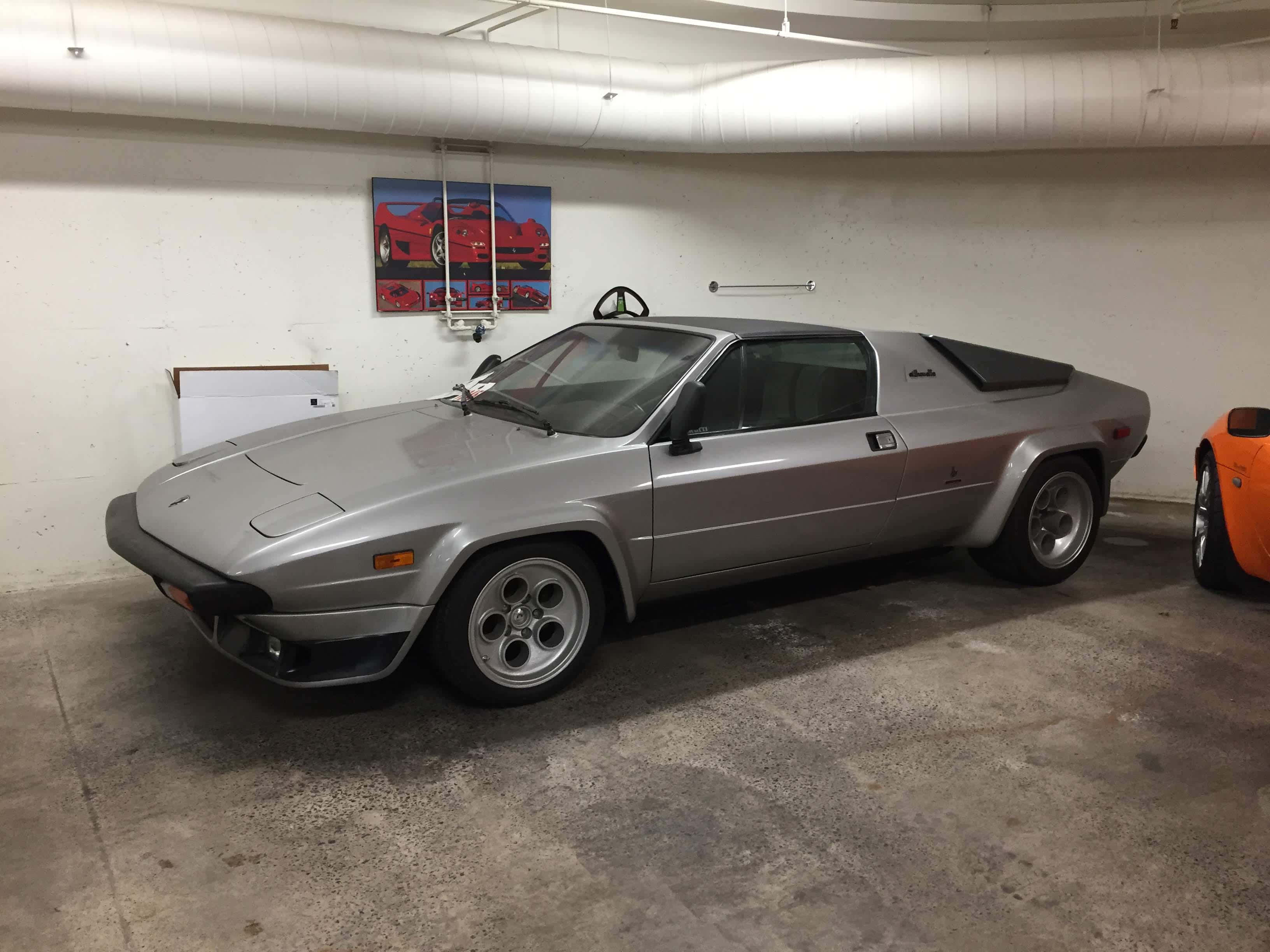 Lamborghini Silhouette 桌布 壁紙 — 一輛時尚的銀色DeLorean跑車停放在混凝土車庫中，車庫有外露管道和牆上的紅色汽車海報。