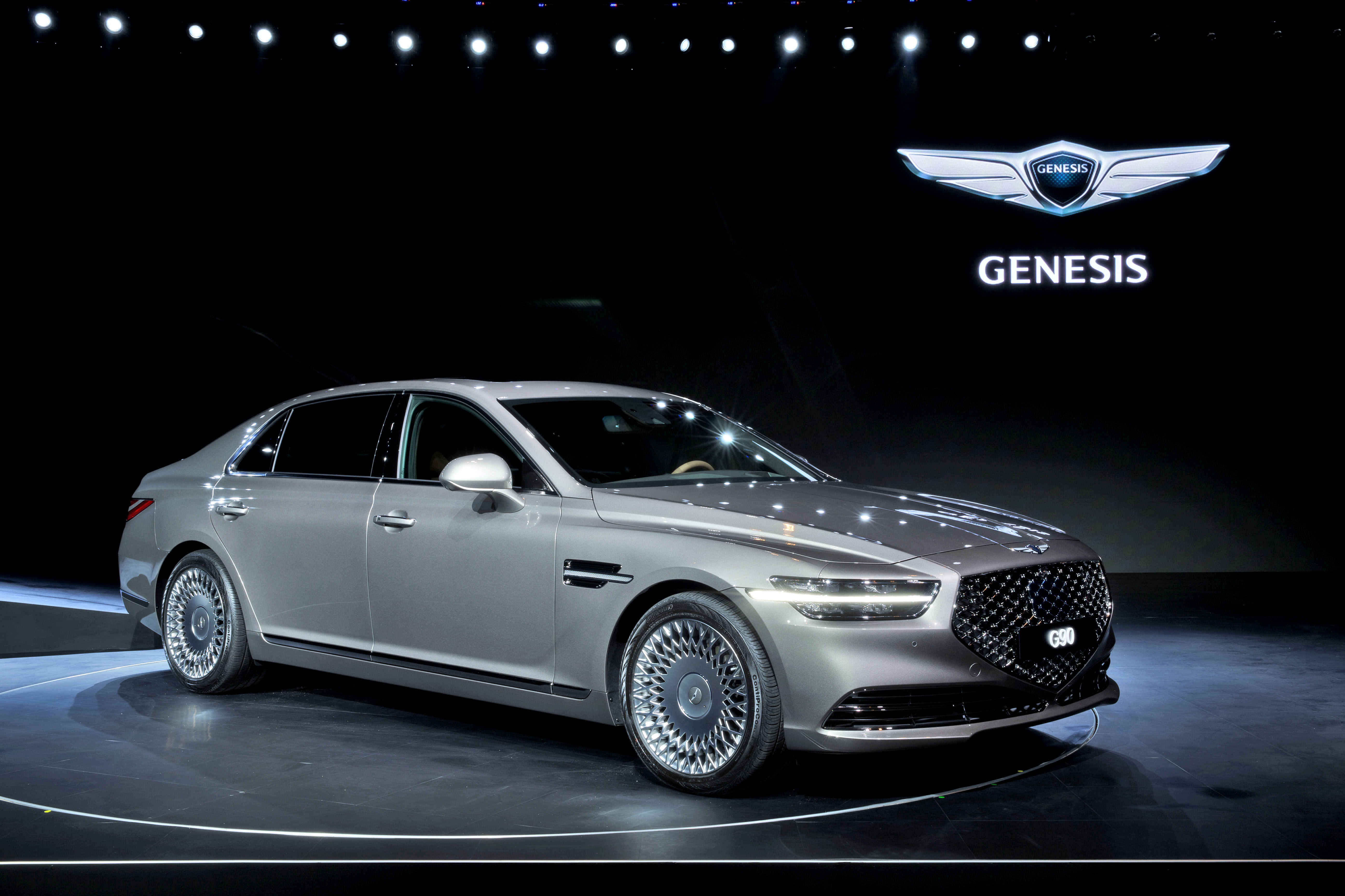 創世紀 桌布 壁紙 — 一輛時尚的銀色Genesis G90豪華轎車在黑暗展廳地板上明亮的舞台燈光下閃閃發光。