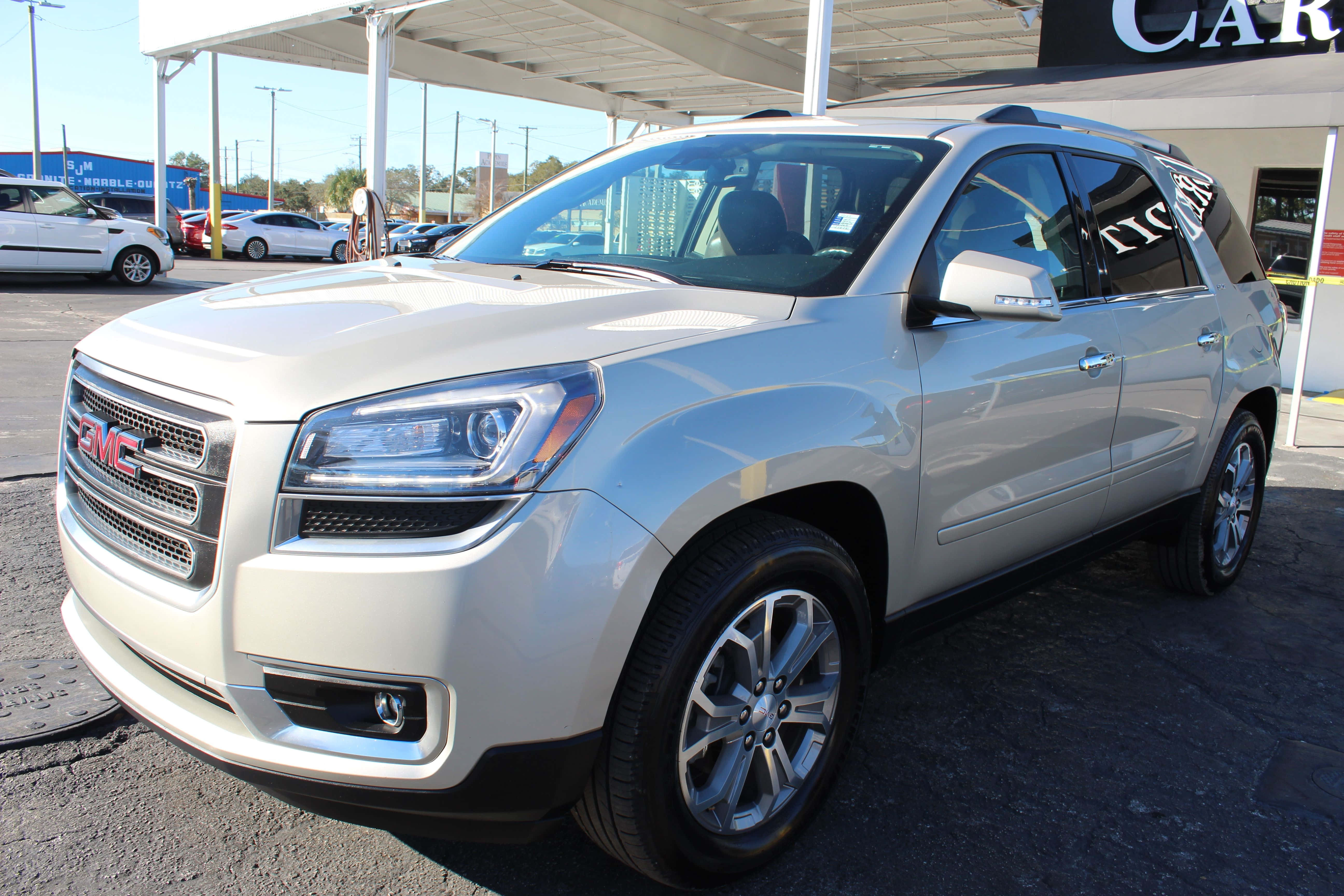 Gmc Acadia 桌布 壁紙 — 一輛時尚的銀色GMC Acadia SUV在明亮的日光下，在汽車經銷商場地中閃閃發光，背景有其他車輛和遮陽篷。