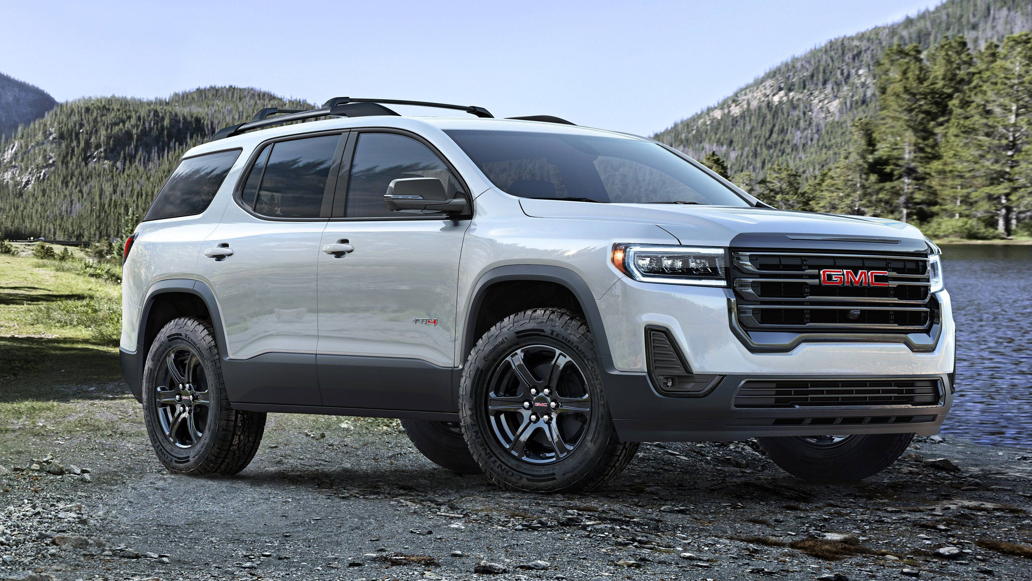 Gmc 桌布 壁紙 — 一輛時尚的銀色GMC SUV，帶有黑色飾邊，停在平靜湖泊旁的礫石岸邊，背景是森林覆蓋的山脈。