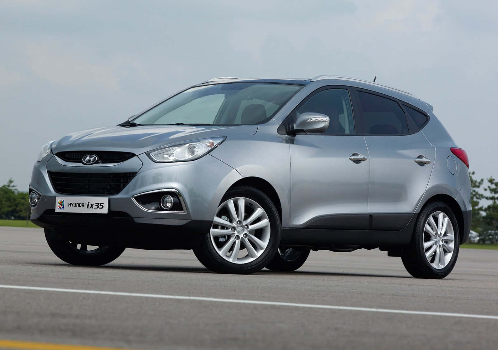 Hyundai 桌布 壁紙 — 一輛時尚的銀色Hyundai ix35 SUV停放在瀝青停車場上，天空多雲，背景有綠樹。