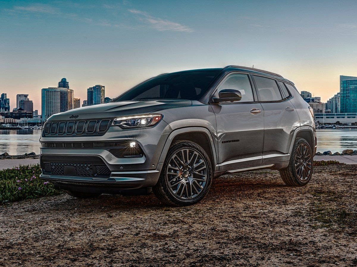 日落 桌布 壁紙 — 一輛時尚的銀色Jeep Compass停泊在泥濘的岸邊，背後是城市天際線和水面，正值黃金時刻。