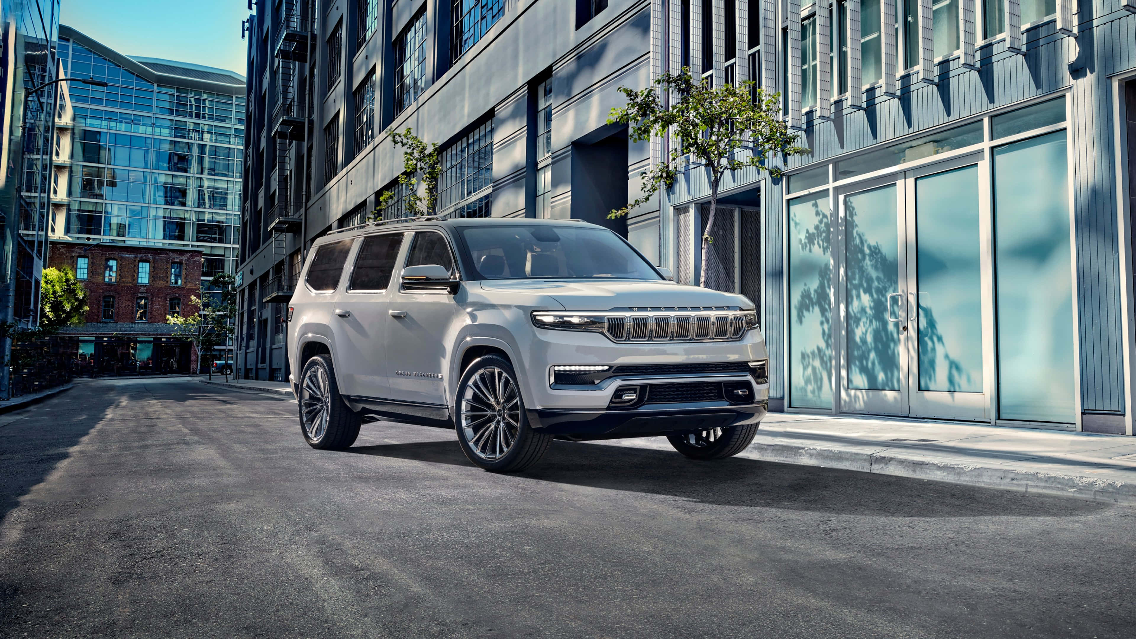 Jeep Wagoneer 桌布 壁紙 — 一輛時尚的銀色Jeep Grand Wagoneer停放在現代城市街道上，兩旁是玻璃和鋼鐵建築，在明亮的日光下。