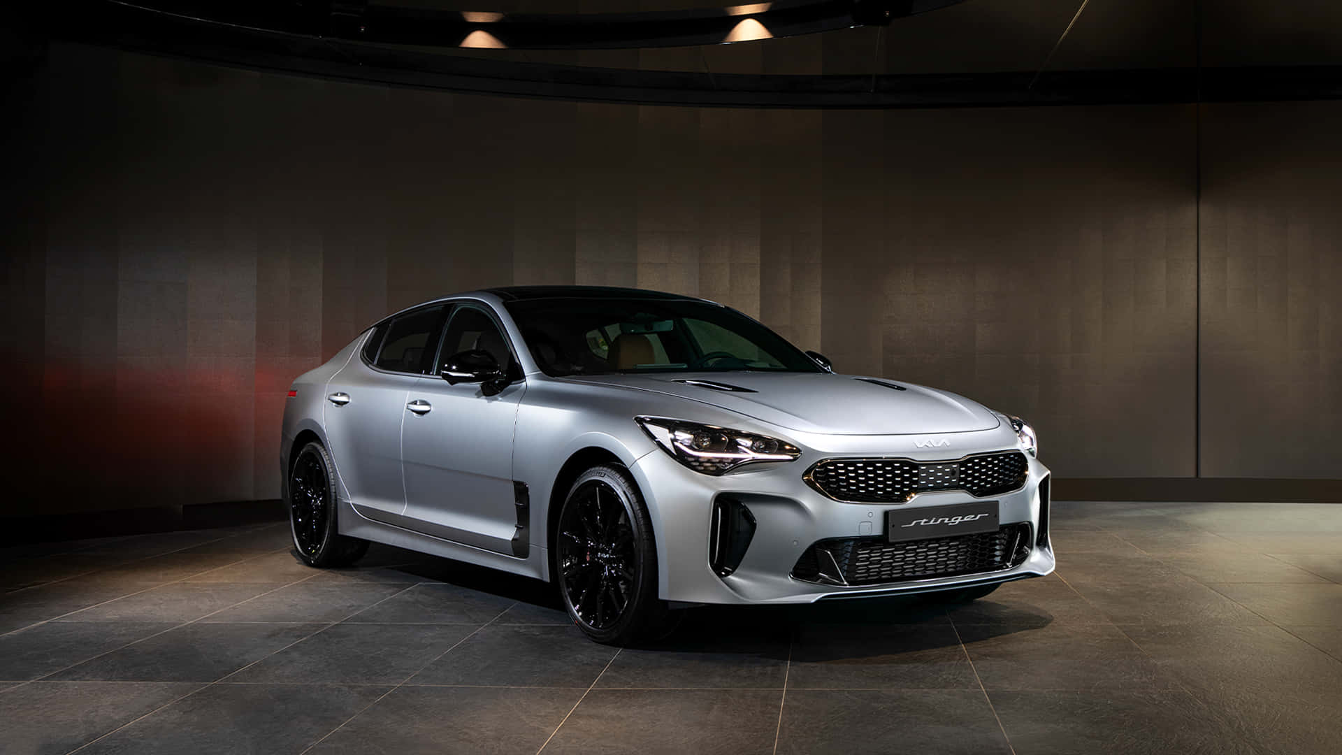 Kia Stinger 桌布 壁紙 — 一輛時尚的銀色Kia Stinger運動轎車在戲劇性的工作室燈光下，以深色紋理牆壁為背景展示。