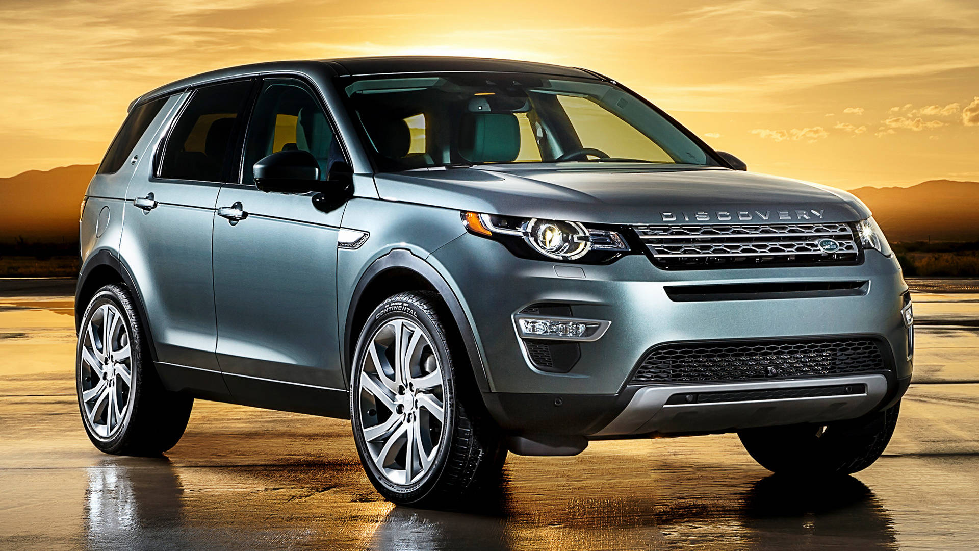 land rover iPhone 桌布 壁紙 — 一輛時尚的銀色Land Rover Discovery SUV在金色夕陽下閃閃發光，在潮濕的沙漠道路上投下長長的影子。