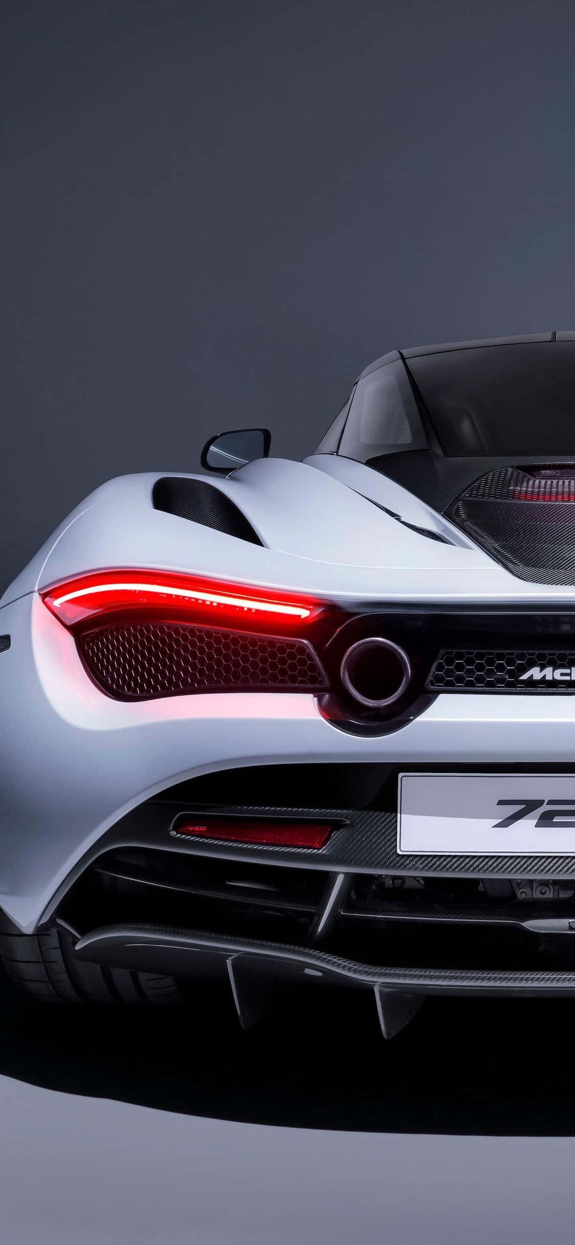 iPhone Xs Mclaren 720s 桌布 壁紙 — 一輛時尚的銀色McLaren 720S跑車，帶有發光的紅色尾燈和碳纖維飾邊，背景為深色工作室。
