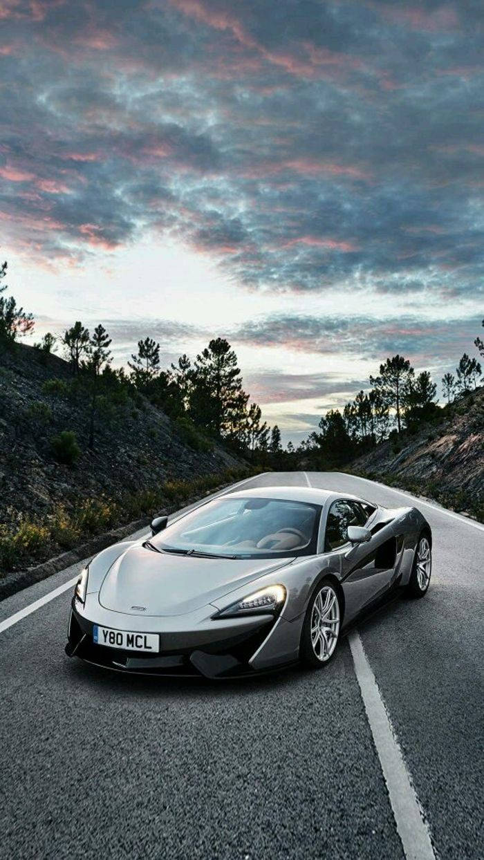 桌布 壁紙 — 一輛時尚的銀色McLaren跑車沿著蜿蜒的山路行駛，在戲劇性的暮色天空下，天空被粉紅色和紫色的雲彩塗抹。