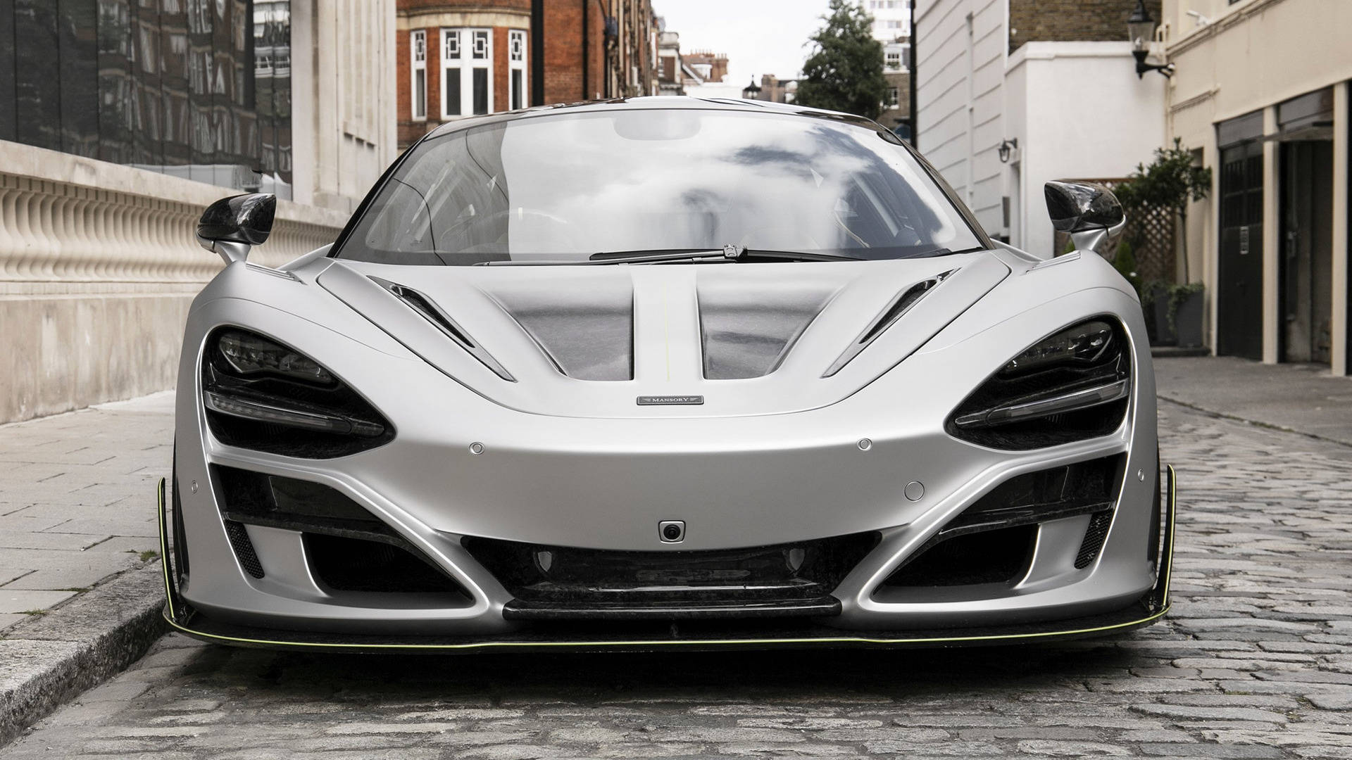 mclaren 720s 4K 桌布 壁紙 — 一輛時尚的銀色McLaren跑車，帶有黃色點綴，停泊在都市建築物之間的鵝卵石街道上。