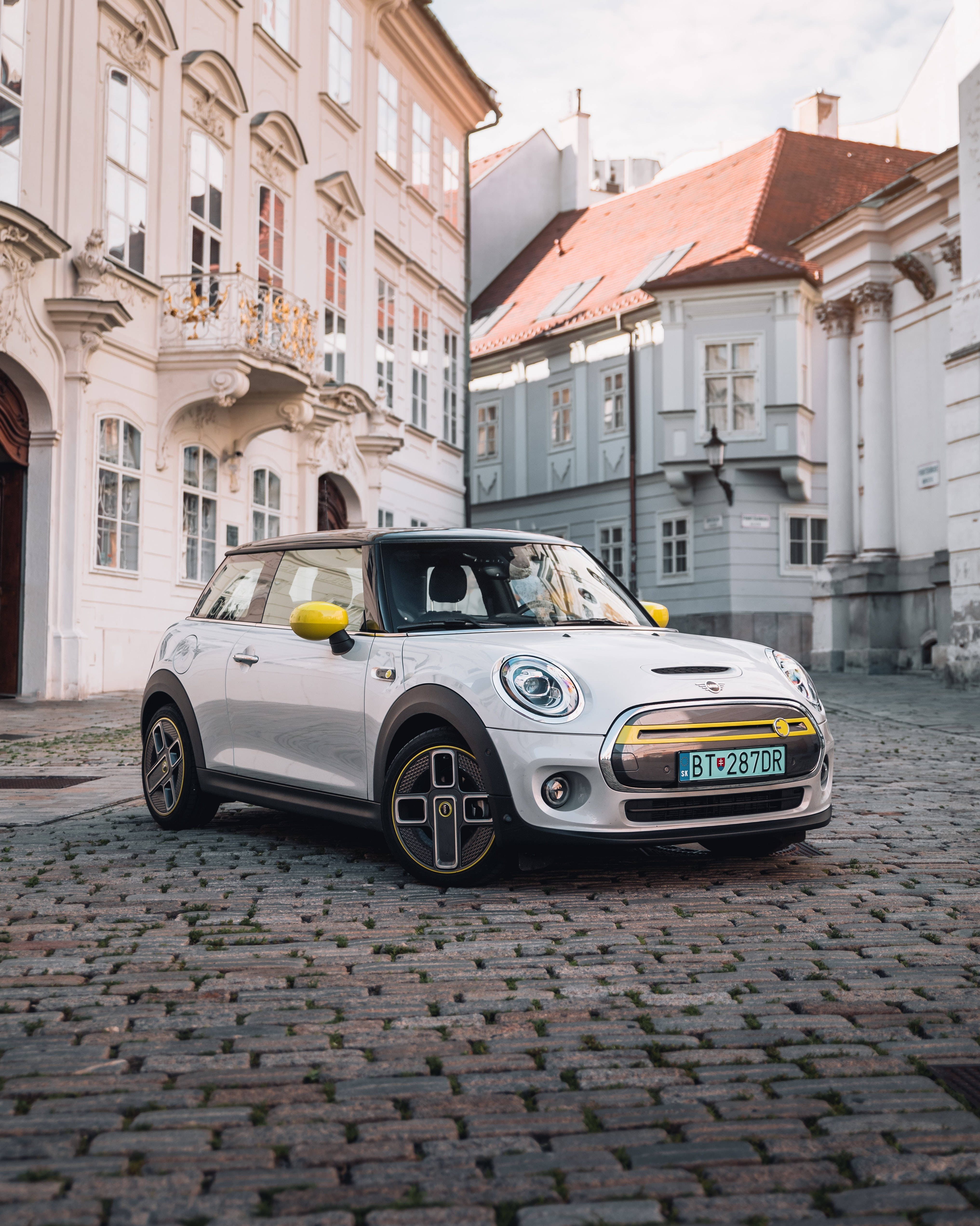 Mini Cooper 桌布 壁紙 — 一輛時尚的銀色MINI Cooper帶有黃色點綴，停在鵝卵石街道上，兩旁是華麗的歐洲建築。