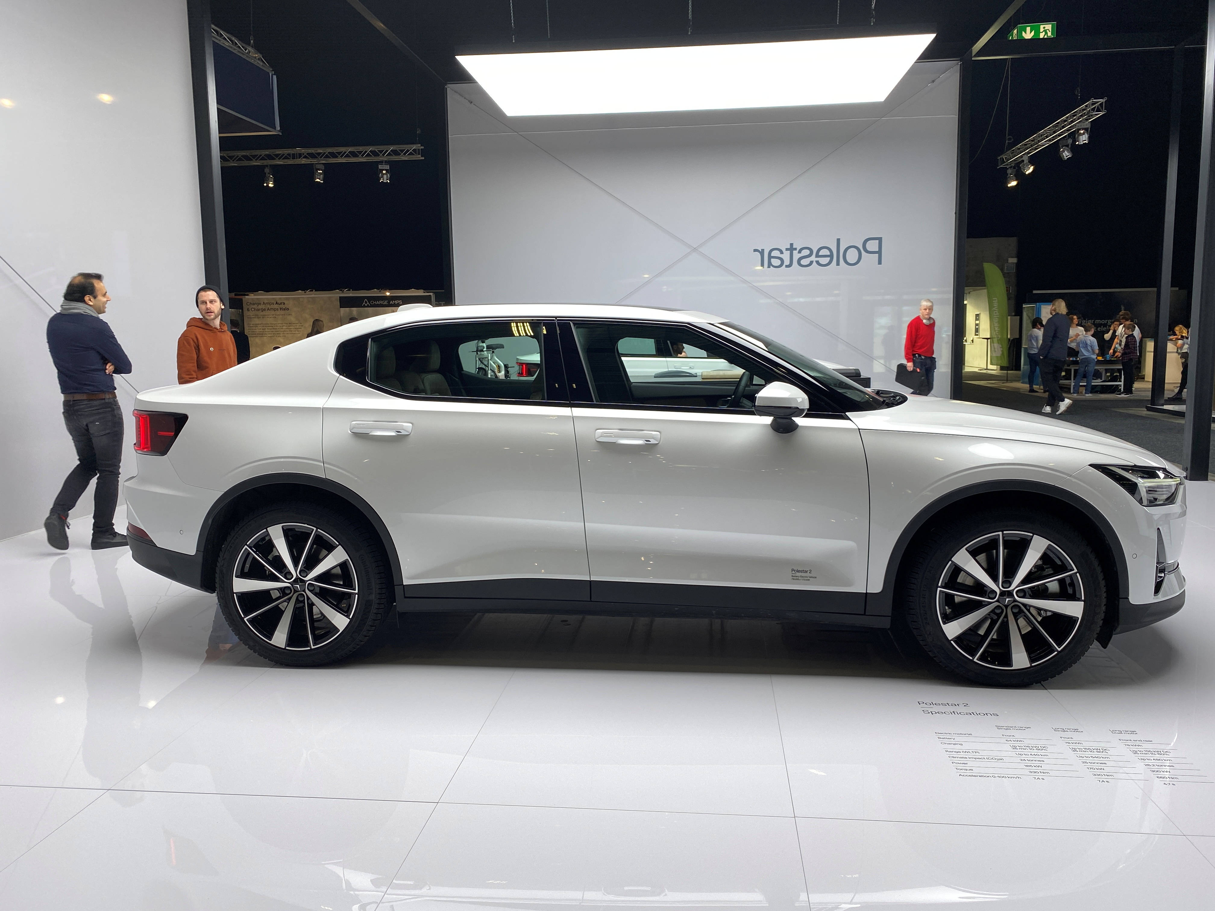 豪華車 桌布 壁紙 — 一輛時尚的銀色Polestar電動車在汽車展覽會上，在光亮的白色地板上展示，人們在旁觀看。
