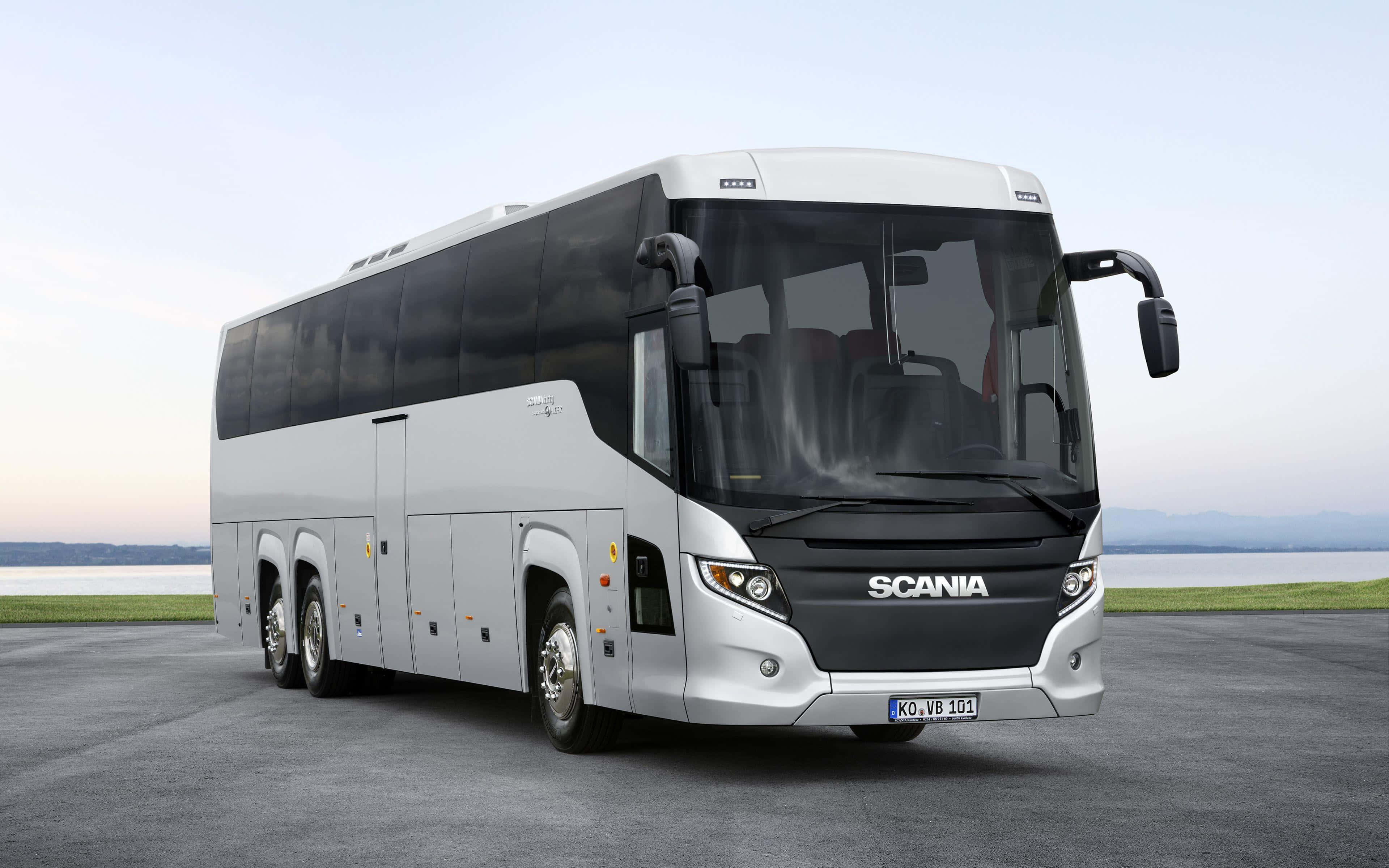 Bus 桌布 壁紙 — 一輛時尚的銀色Scania長途巴士停在近水的瀝青路上，遠處有山巒，天空柔和暮色。