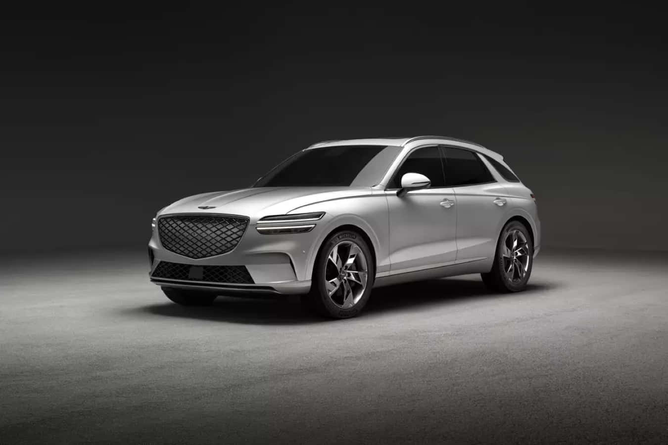 Genesis Gv70 桌布 壁紙 — 一輛時尚的銀色SUV，配備現代水箱護罩和銳利線條，在戲劇性的攝影棚燈光下，以深色漸層背景展示。