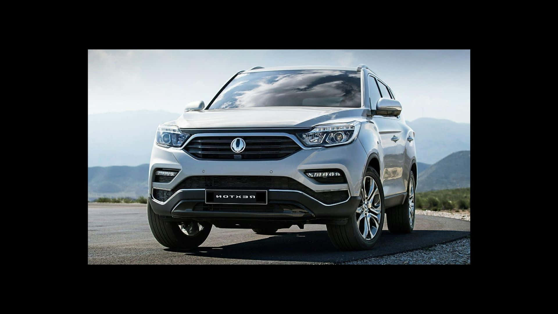 Ssangyong 桌布 壁紙 — 一輛時尚的銀色SUV停在鋪砌的道路上，背景是山區地形，天空明亮。