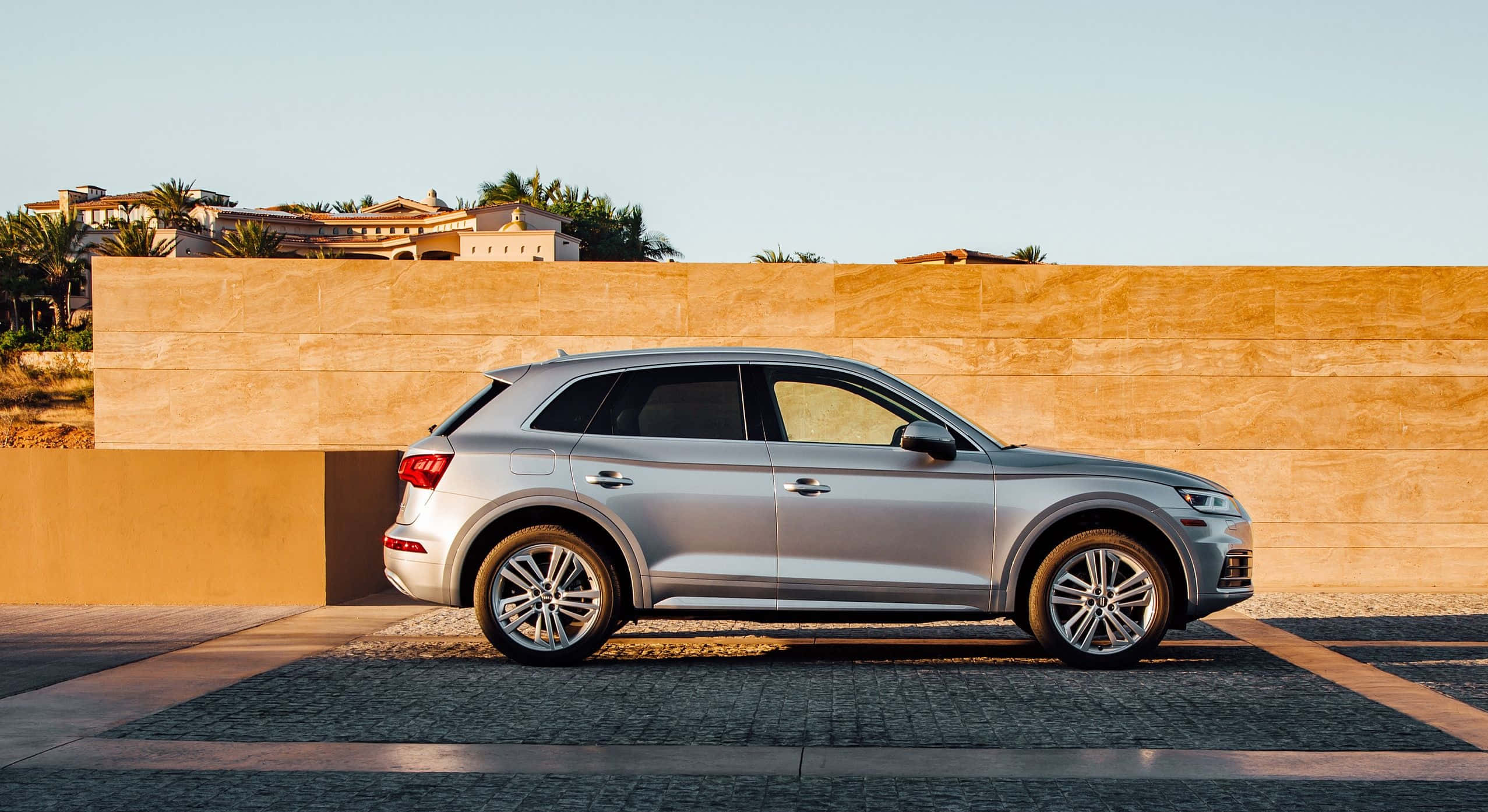 Audi Sq5 桌布 壁紙 — 一輛時尚的銀色SUV停放在暖色調的石牆旁，天空晴朗，遠處有棕櫚樹和房屋。