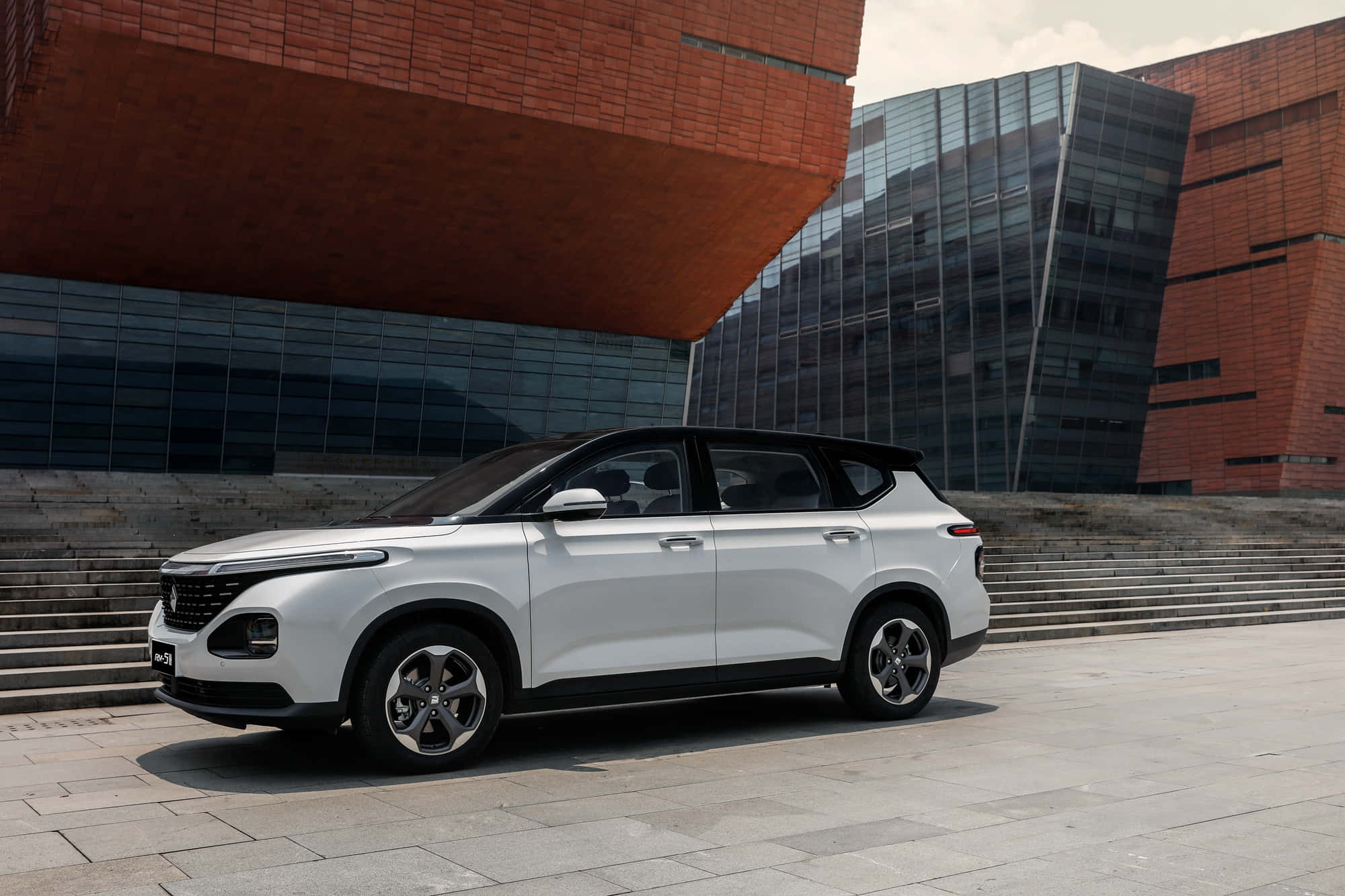 Baojun 桌布 壁紙 — 一輛時尚的銀色SUV停放在石頭廣場上，前方是一座現代建築，擁有醒目的紅磚和玻璃外牆。
