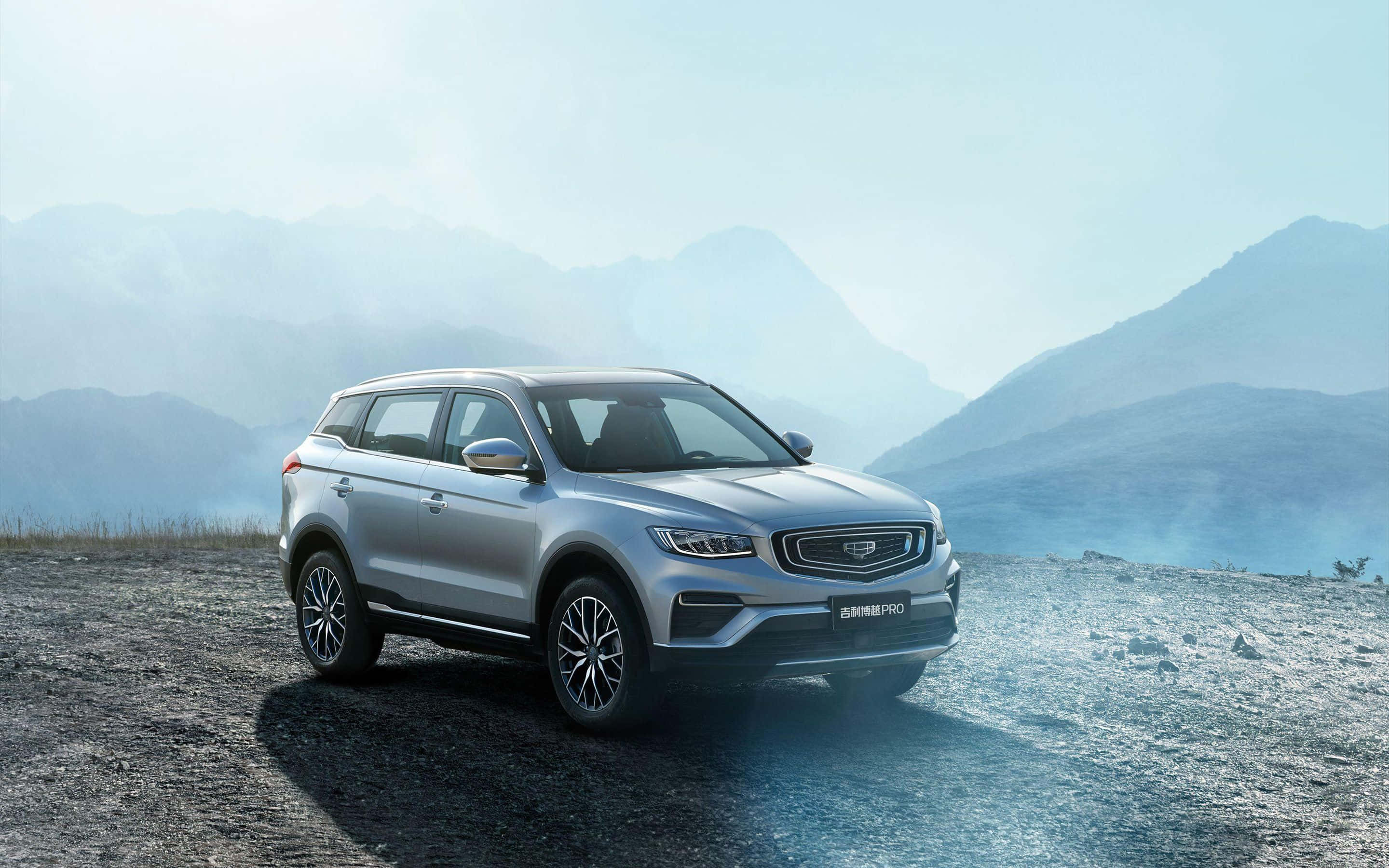Geely 桌布 壁紙 — 一輛時尚的銀色SUV停泊在崎嶇的山景瞭望點，背景是霧氣繚繞的山峰，天空柔和湛藍。