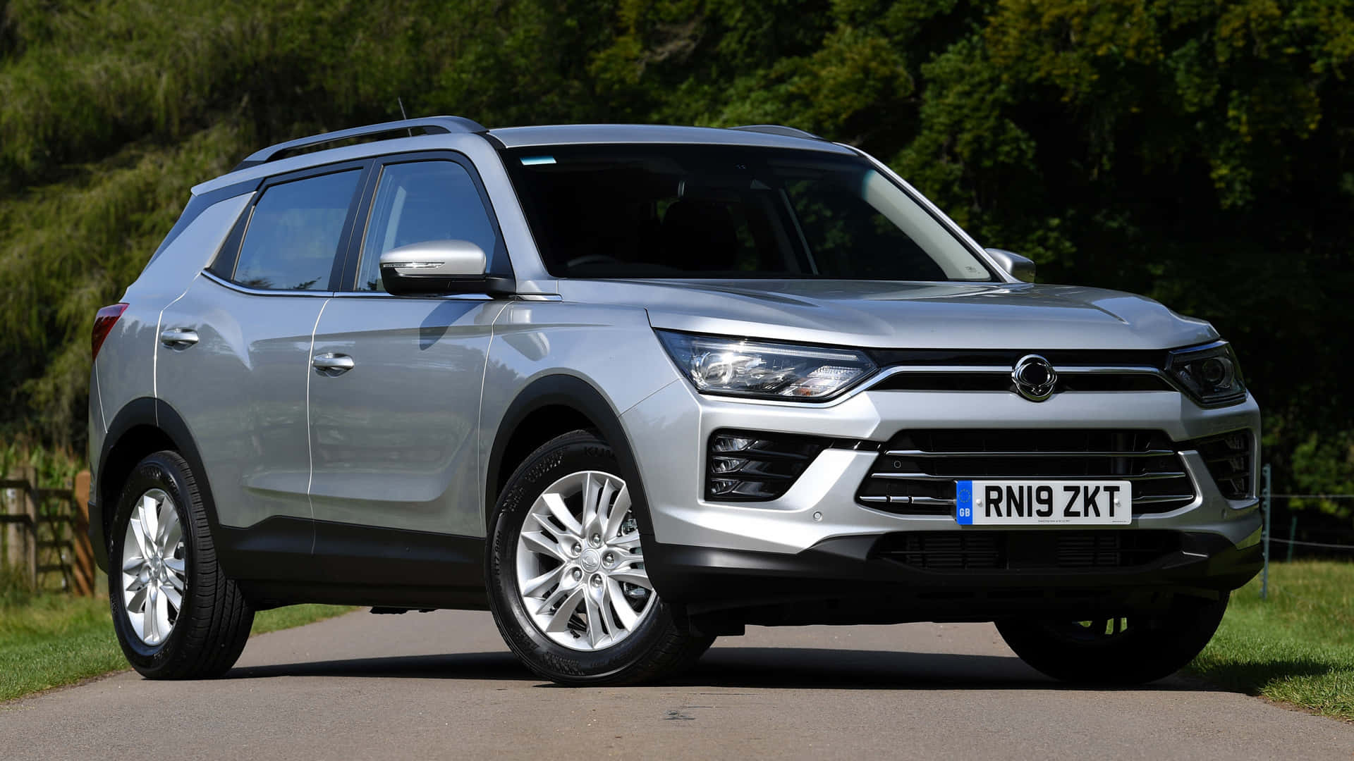 Ssangyong 桌布 壁紙 — 一輛時尚的銀色SUV停泊在鋪砌的道路上，背景有茂密的綠樹和草地，在明亮的日光下。