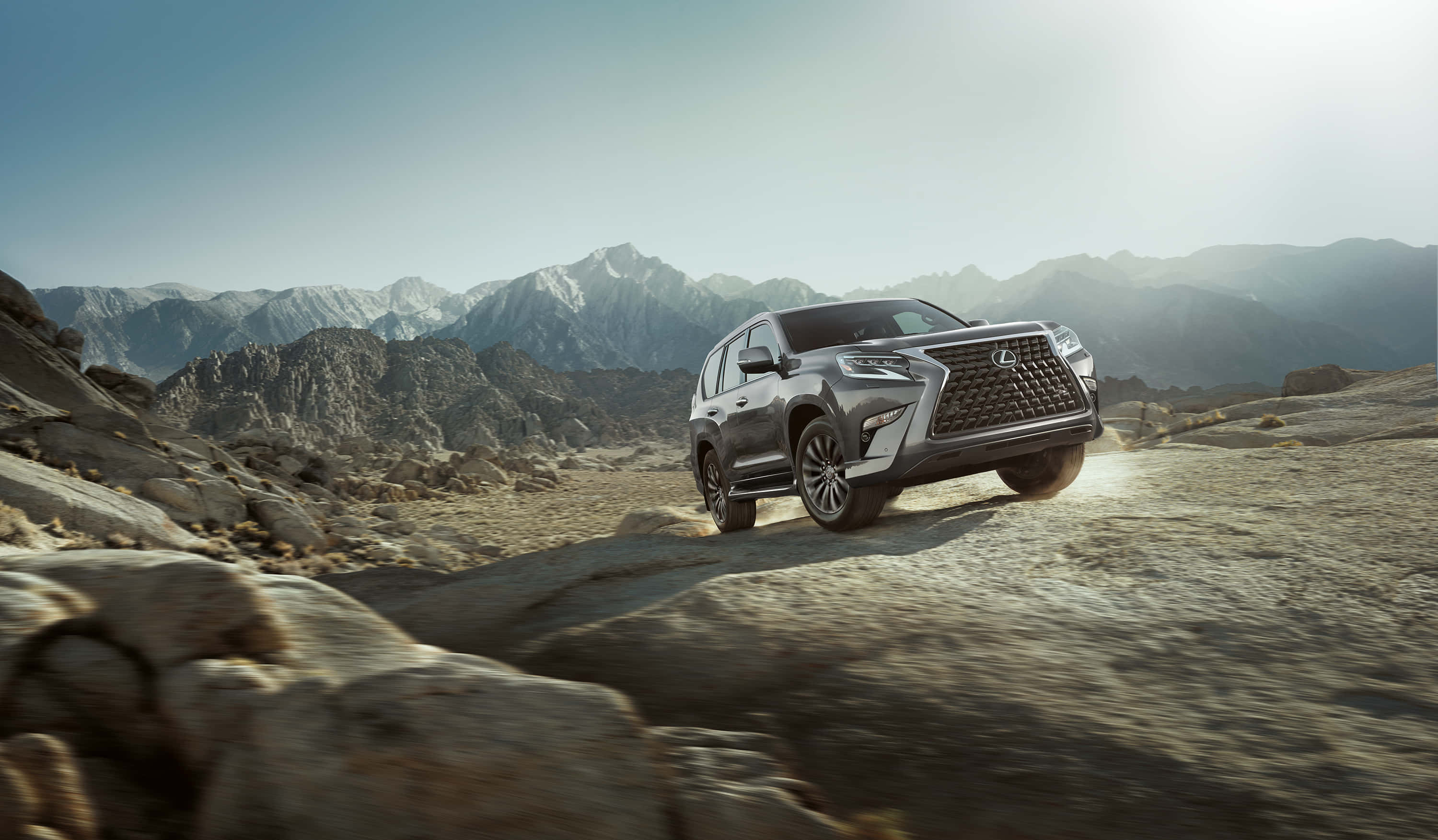 Lexus Gx 460 桌布 壁紙 — 一輛時尚的銀色SUV在明亮的陽光下征服崎嶇的山地地形，遠處是白雪覆蓋的山峰。