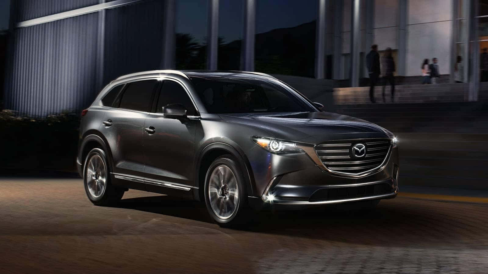 Mazda Cx-9 桌布 壁紙 — 一輛時尚的銀色SUV在都市夜景中滑行，車頭燈亮起，背景是現代建築。