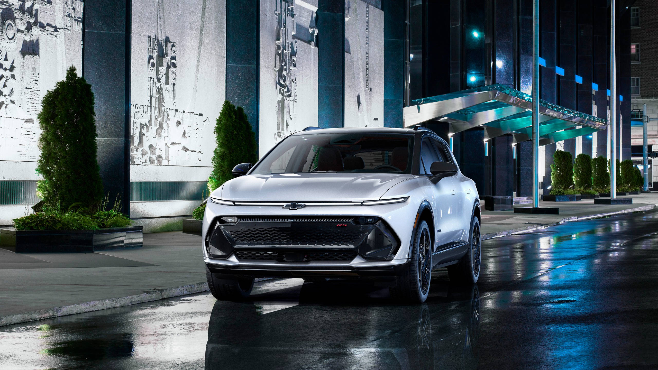 Chevrolet 桌布 壁紙 — 一輛時尚的銀色SUV在霓虹燈下閃耀，映照在濕漉漉的城市街道上，反射出周圍建築物的藍色和綠色色調。