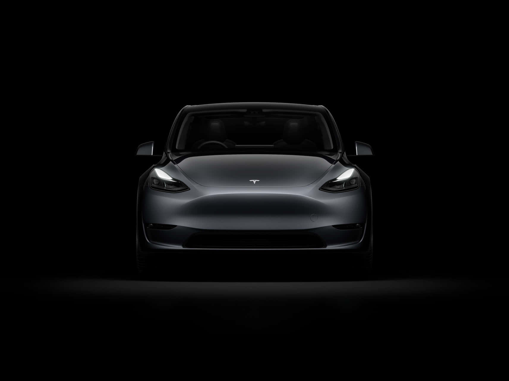 特斯拉 桌布 壁紙 — 一輛時尚的銀色Tesla Model 3正面朝前，車頭燈在漆黑的工作室背景中發光。