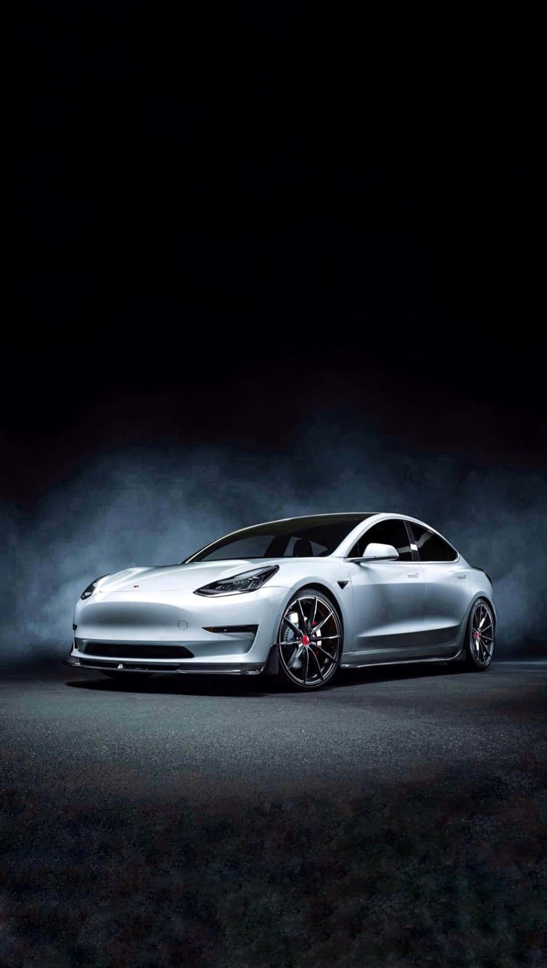 Tesla iPhone 桌布 壁紙 — 一輛時尚的銀色Tesla Model 3在煙霧瀰漫的深色背景下，於戲劇性的聚光燈下閃耀。