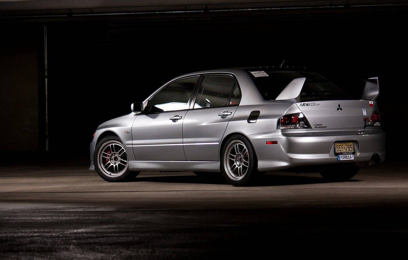 Mitsubishi 桌布 壁紙 — 一輛時尚的銀色三菱Lancer Evolution停放在燈光昏暗的車庫中，戲劇性的聚光燈凸顯其運動設計。