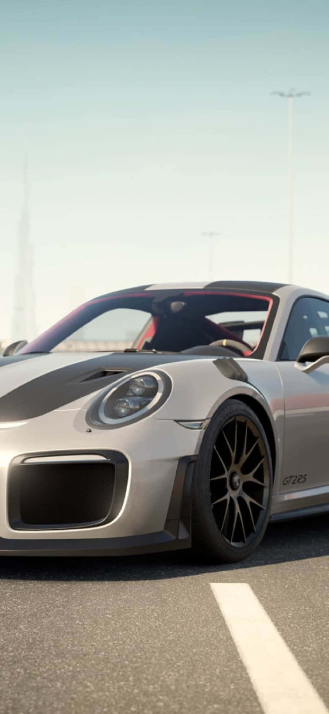 保時捷 911 桌布 壁紙 — 一輛時尚的銀色保時捷911 GT2 RS，帶有黑色飾邊和紅色內飾，停在陽光明媚的道路上，遠處是城市天際線。