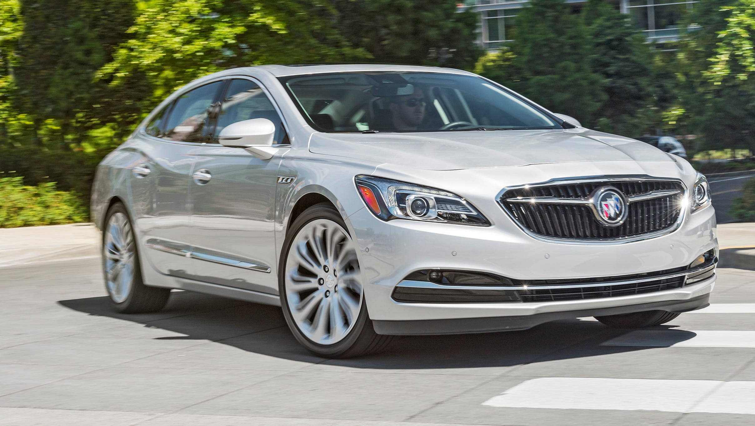 Buick 桌布 壁紙 — 一輛時尚的銀色別克轎車在陽光明媚的城市道路上平穩行駛，周圍環繞著茂密的綠樹。