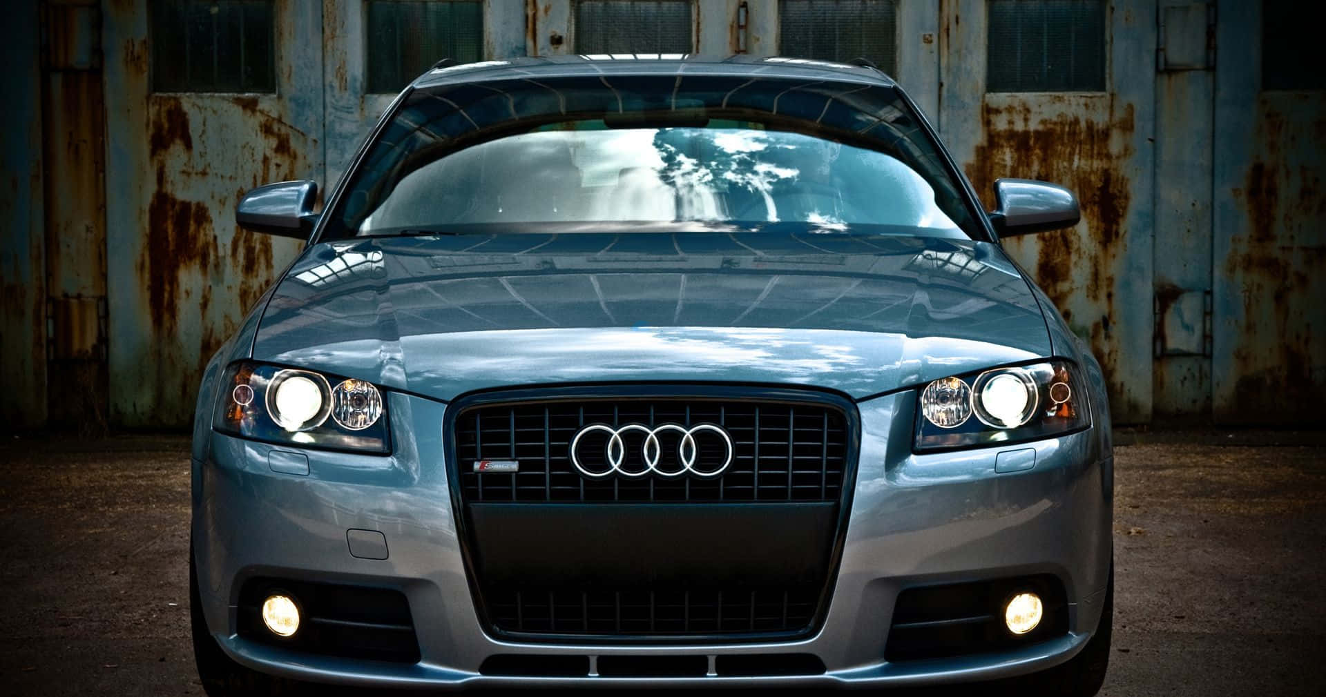 Audi A3 桌布 壁紙 — 一輛時尚的銀色奧迪跑車，車頭燈閃爍，停在生鏽的工業門前。