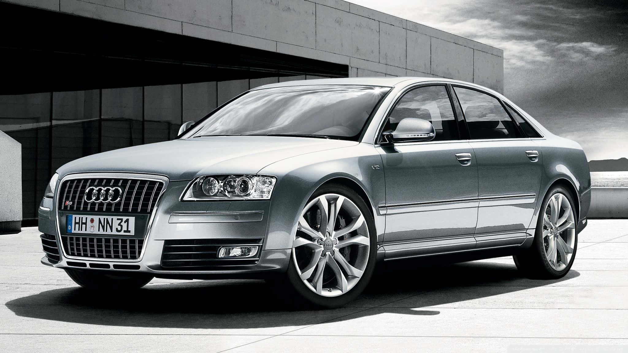 Audi S8 桌布 壁紙 — 一輛時尚的銀色奧迪轎車停放在現代混凝土建築前，背景是戲劇性的多雲天空。