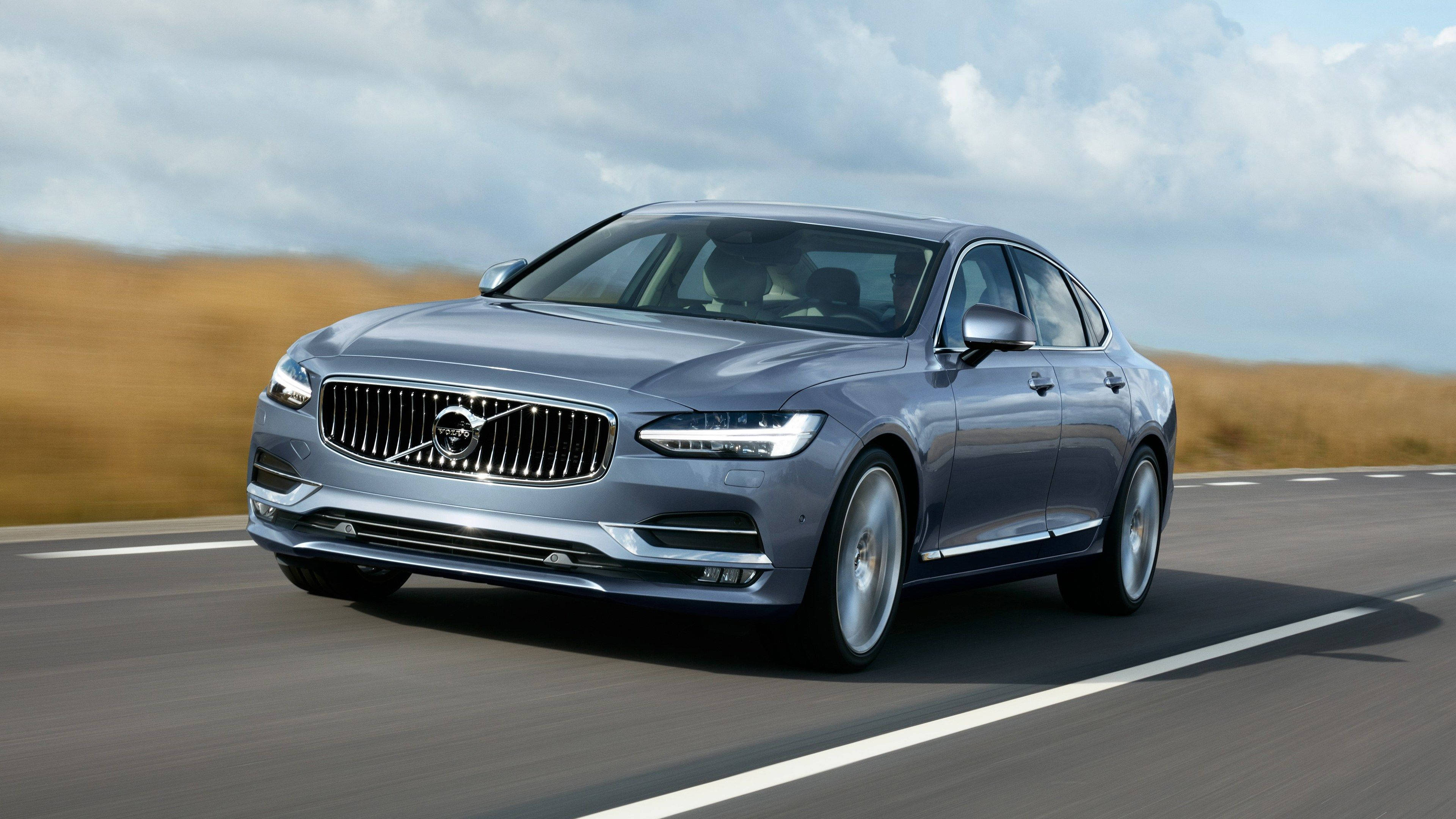 Volvo 桌布 壁紙 — 一輛時尚的銀色富豪房車在陰天下行駛在鋪砌的道路上，兩旁是金色的草地。