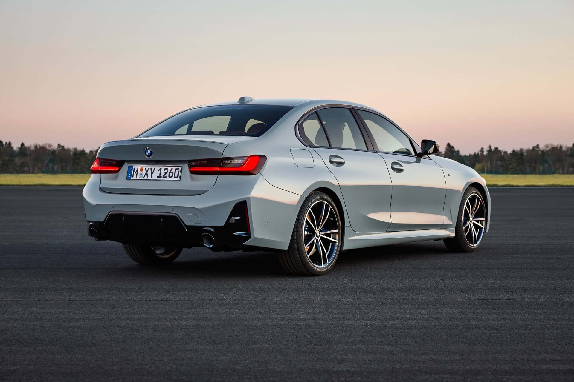 BMW 動圖 桌布 壁紙 — 一輛時尚的銀色寶馬3系轎車停在瀝青路上，背景是日落時分柔和的粉紅色和橙色天空。