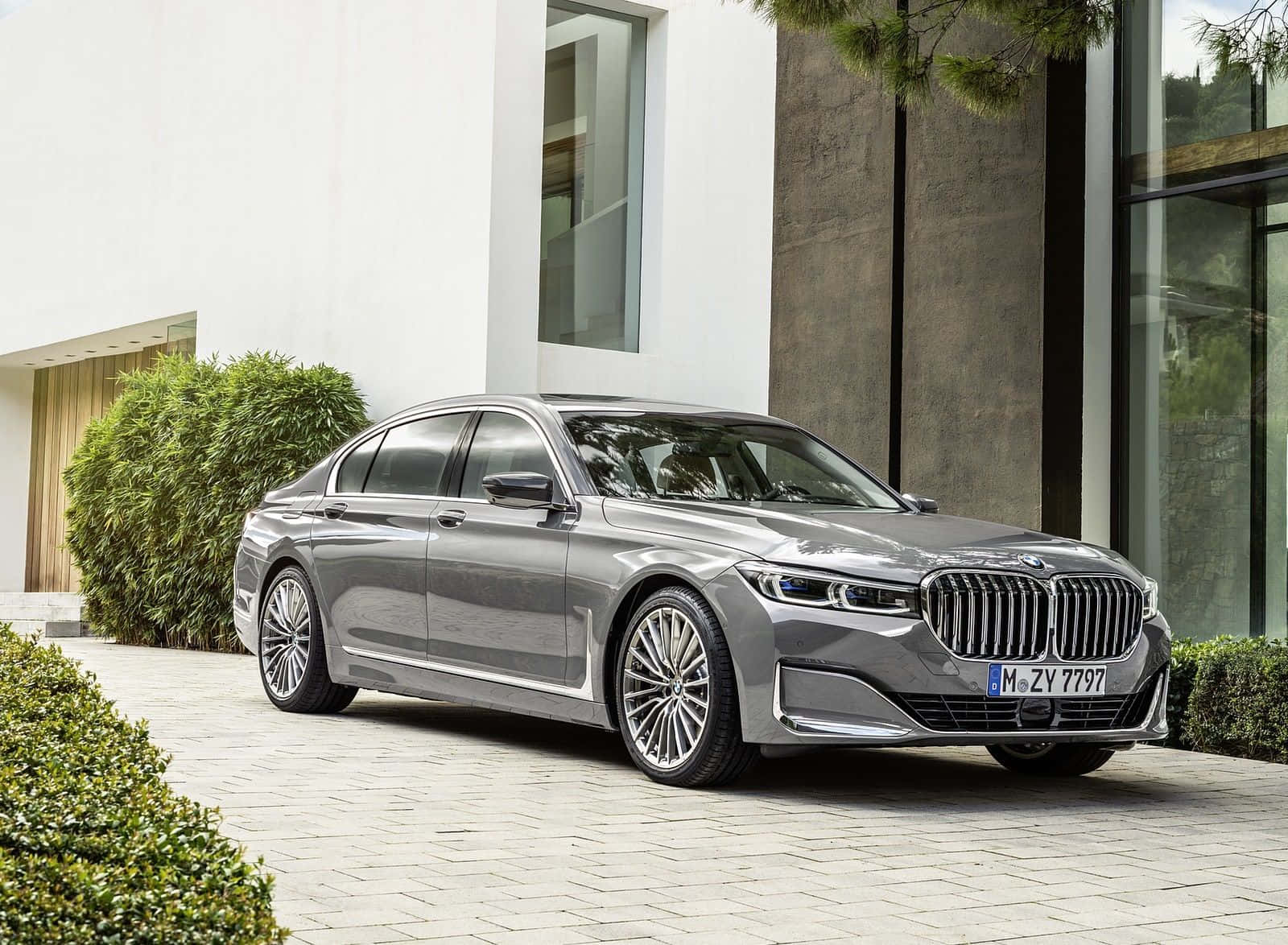 Bmw 7 Series 桌布 壁紙 — 一輛時尚的銀色寶馬7系列轎車停放在石頭車道上，旁邊是一座現代白色和混凝土房屋，周圍有綠色灌木。