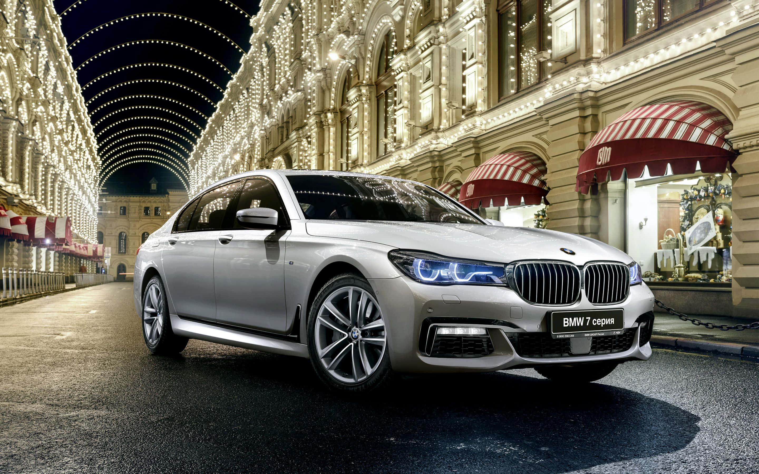 Bmw 7 Series 桌布 壁紙 — 一輛時尚的銀色寶馬7系列轎車在夜晚的歐洲宏偉購物拱廊中，在節日燈串下閃閃發光。