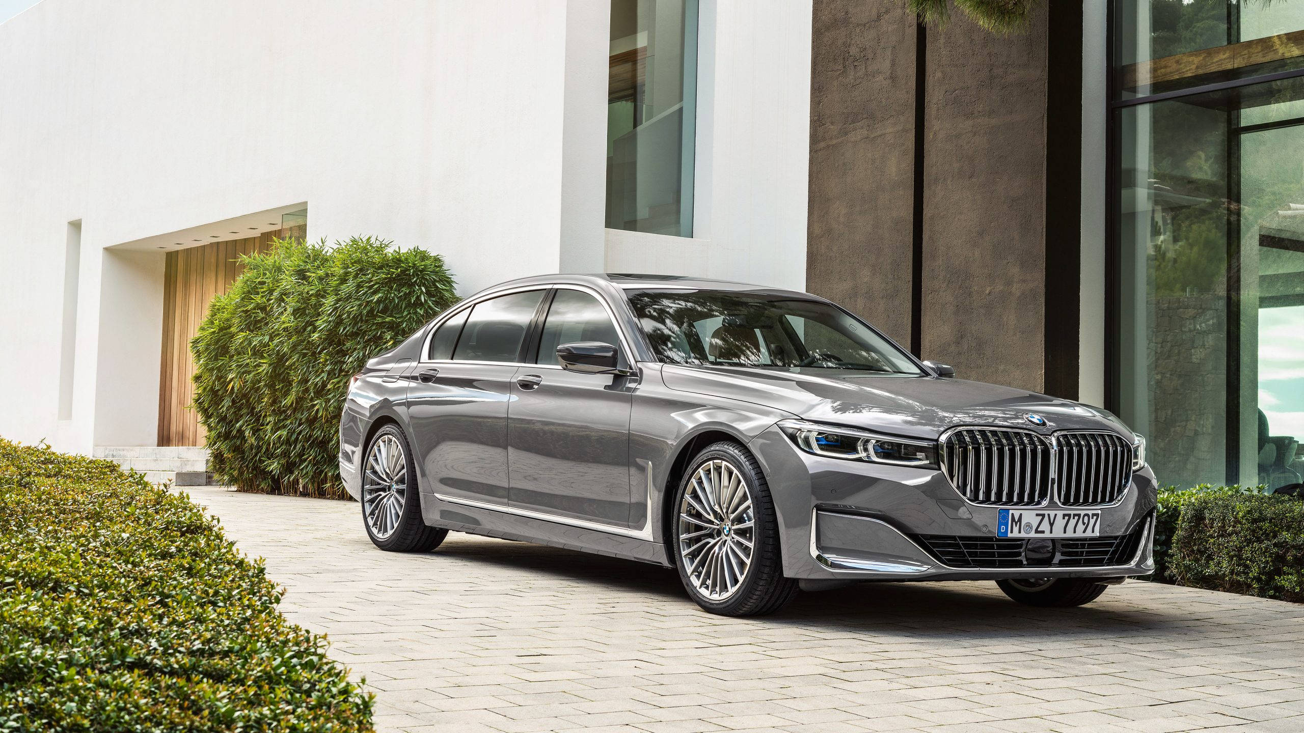 BMW 動圖 桌布 壁紙 — 一輛時尚的銀色寶馬7系轎車停泊在石頭車道上，旁邊是現代建築和修剪整齊的綠色樹籬。